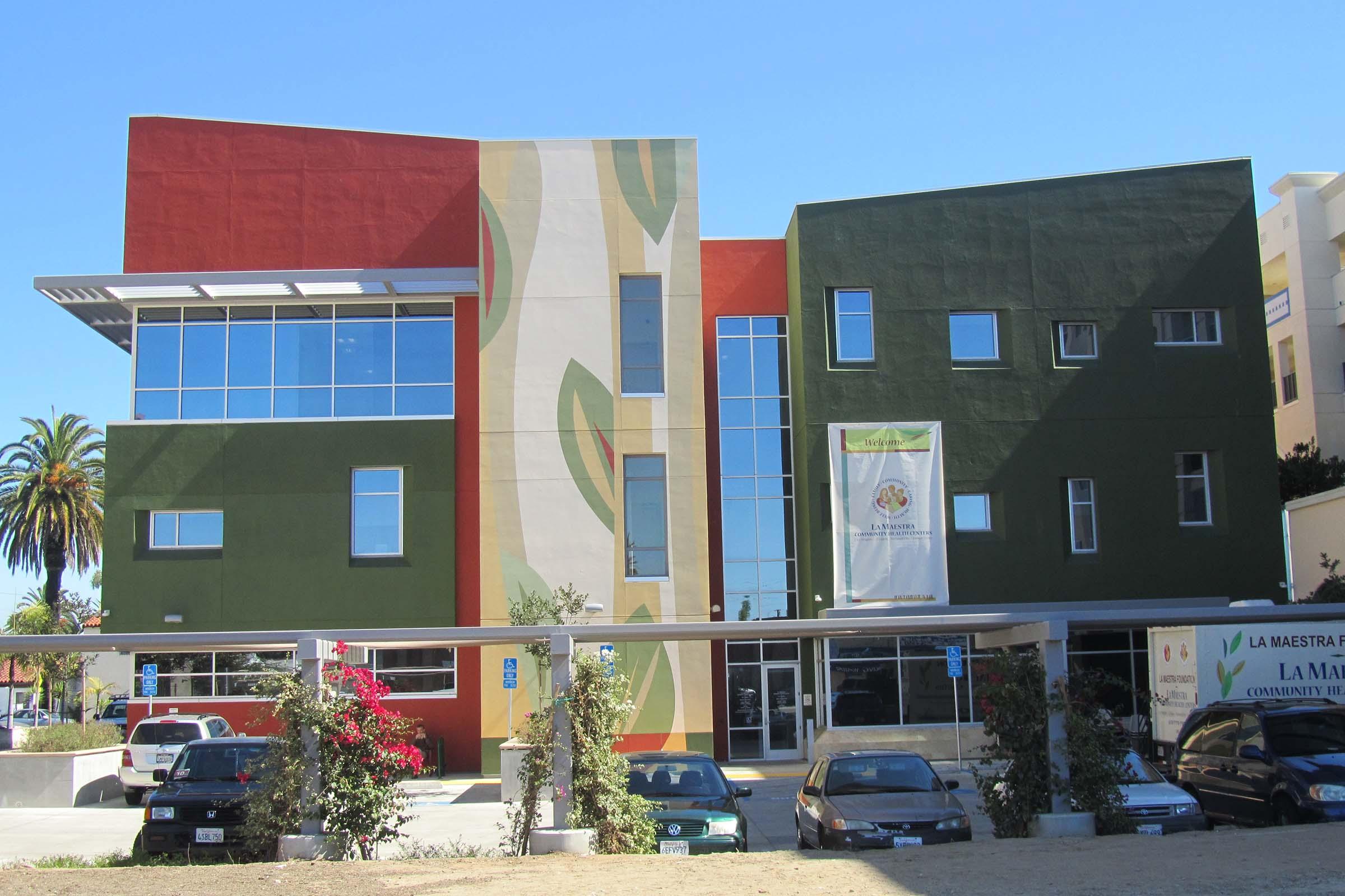 Modern building with a colorful exterior featuring green, red, and white elements. Large windows on the upper floors provide natural light, and a banner hangs at the entrance. In the foreground, several parked cars and landscaped plants add to the setting, with palm trees visible in the background.