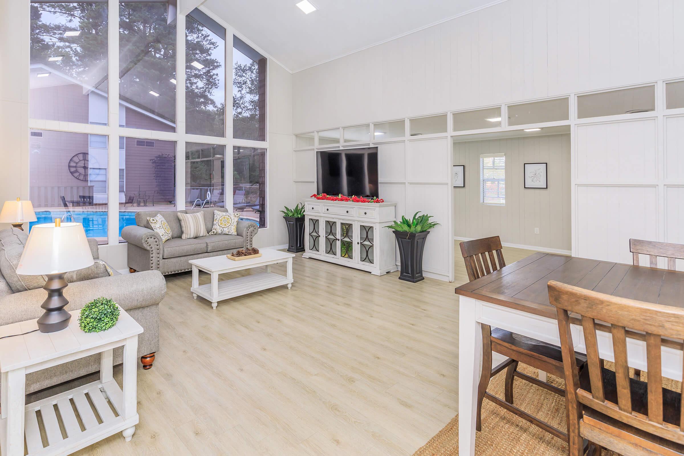 A bright and airy living room featuring large windows, a comfortable gray sofa, a white coffee table, and a TV mounted on the wall. There are decorative plants, a dining table with chairs, and a cozy atmosphere with wood-like flooring. The décor is modern and inviting, with subtle decorative accents.