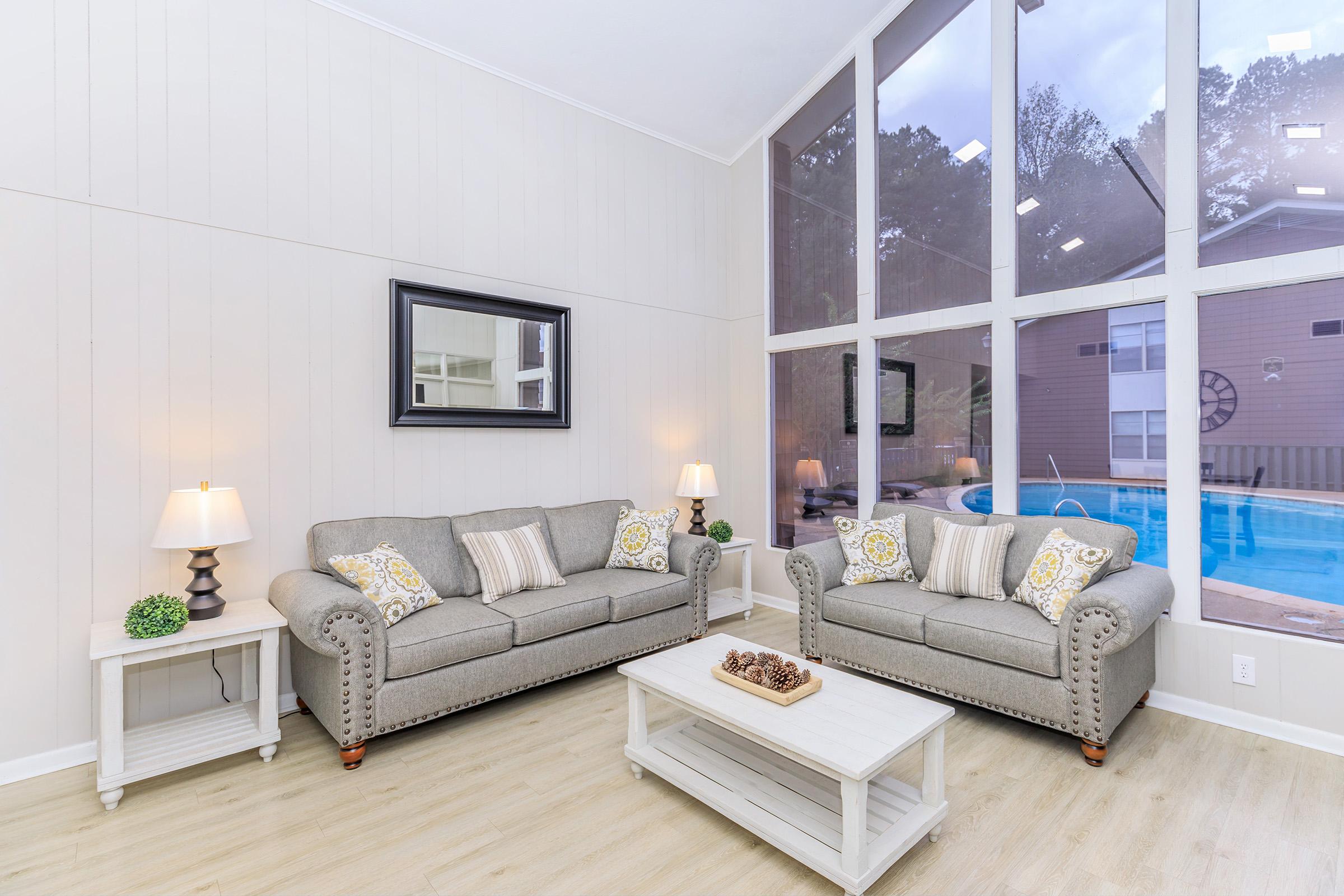 A bright and modern living room featuring two light gray sofas with decorative cushions, a white coffee table, and a stylish wall mirror. Large windows allow natural light to flood the space, revealing a view of a pool outside. Soft lamps on either side add a warm ambiance.