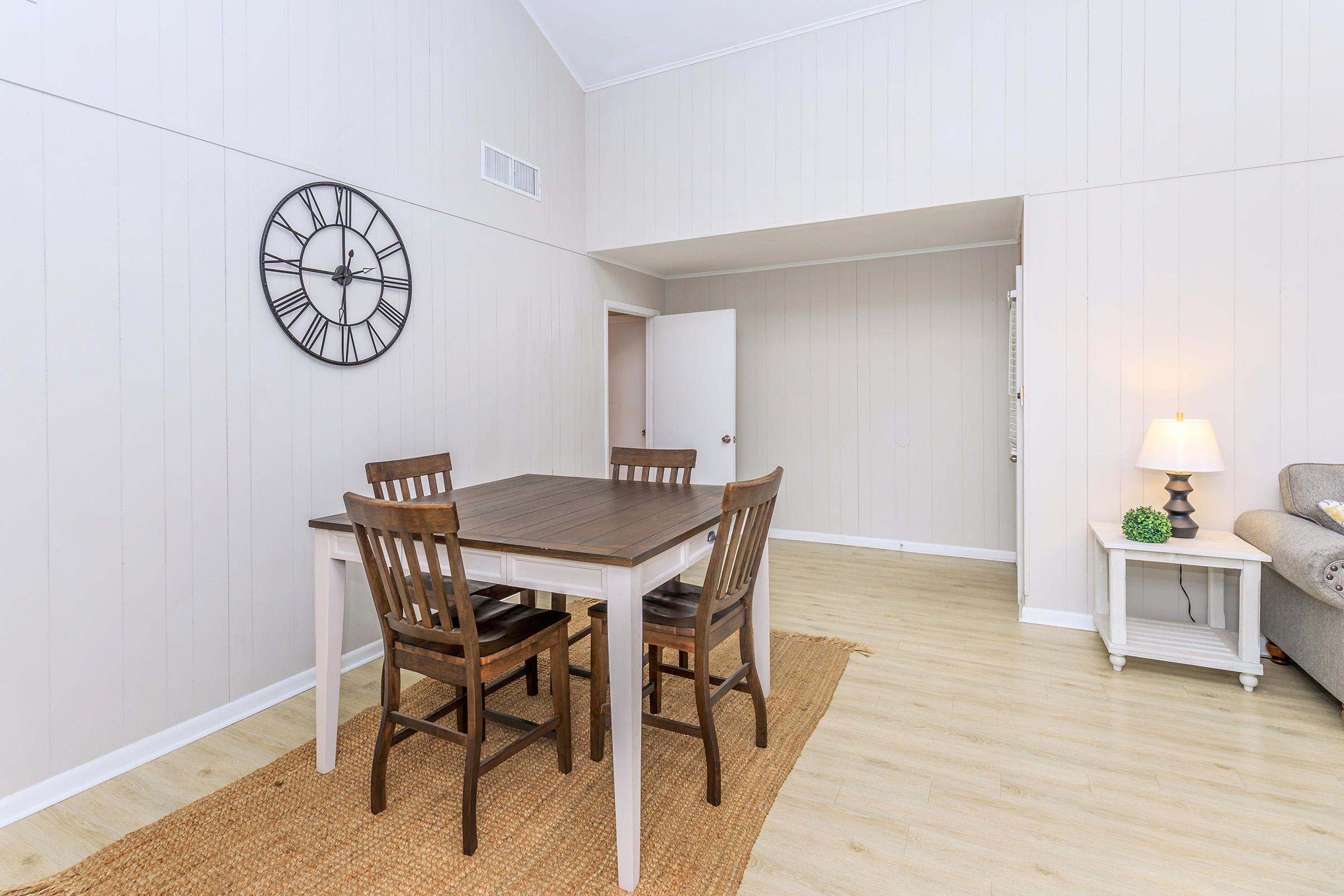 A bright, modern dining area featuring a wooden table with four chairs, a large wall clock, and a lamp on a side table. The walls are light-colored, with a cozy rug on the floor, and there's a doorway leading to another room.