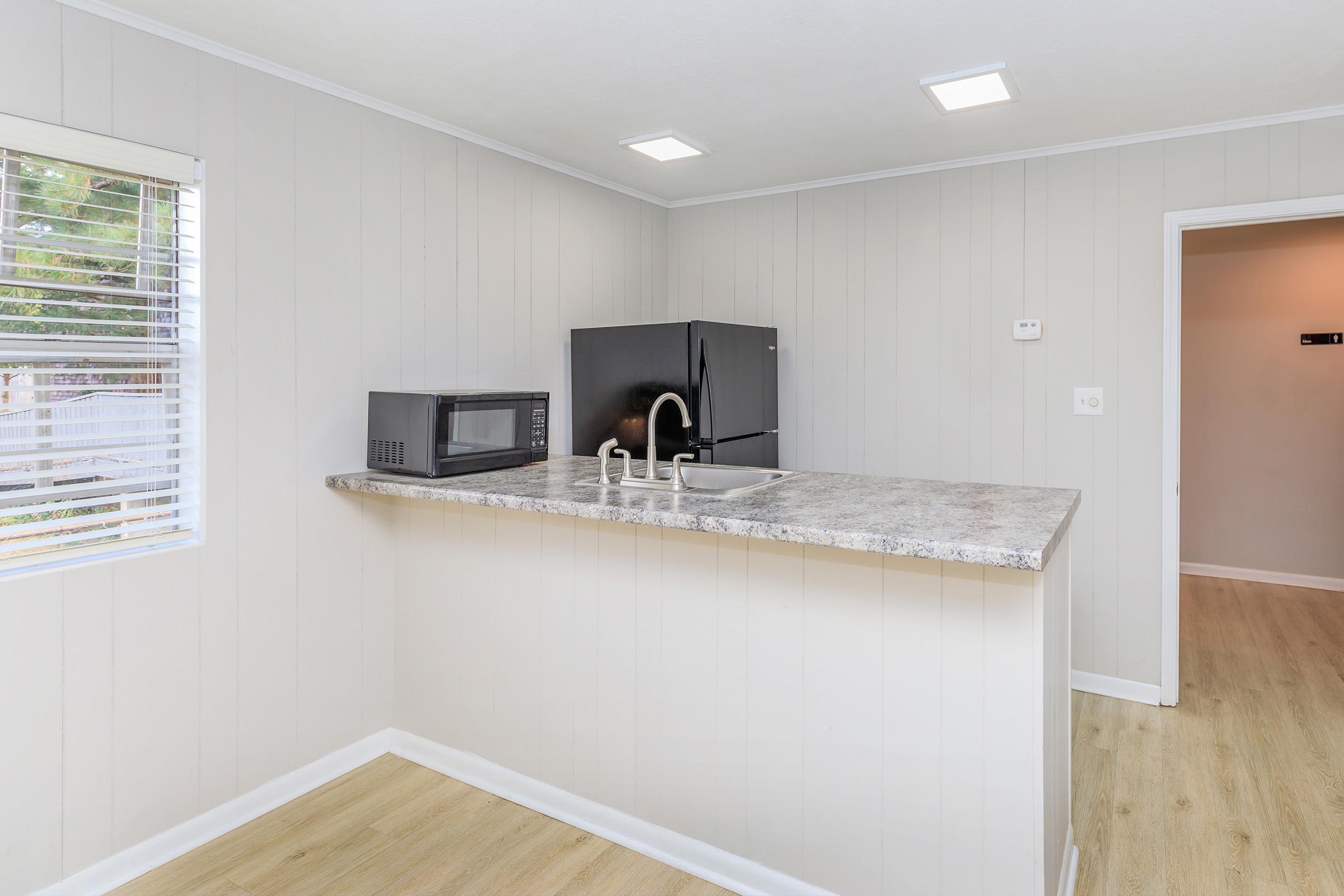 A modern kitchen with light gray walls and vinyl flooring. The room features a countertop with a sink, a black refrigerator, and a microwave on the side. There are bright ceiling lights and a window allowing natural light to enter, creating a clean and airy atmosphere.