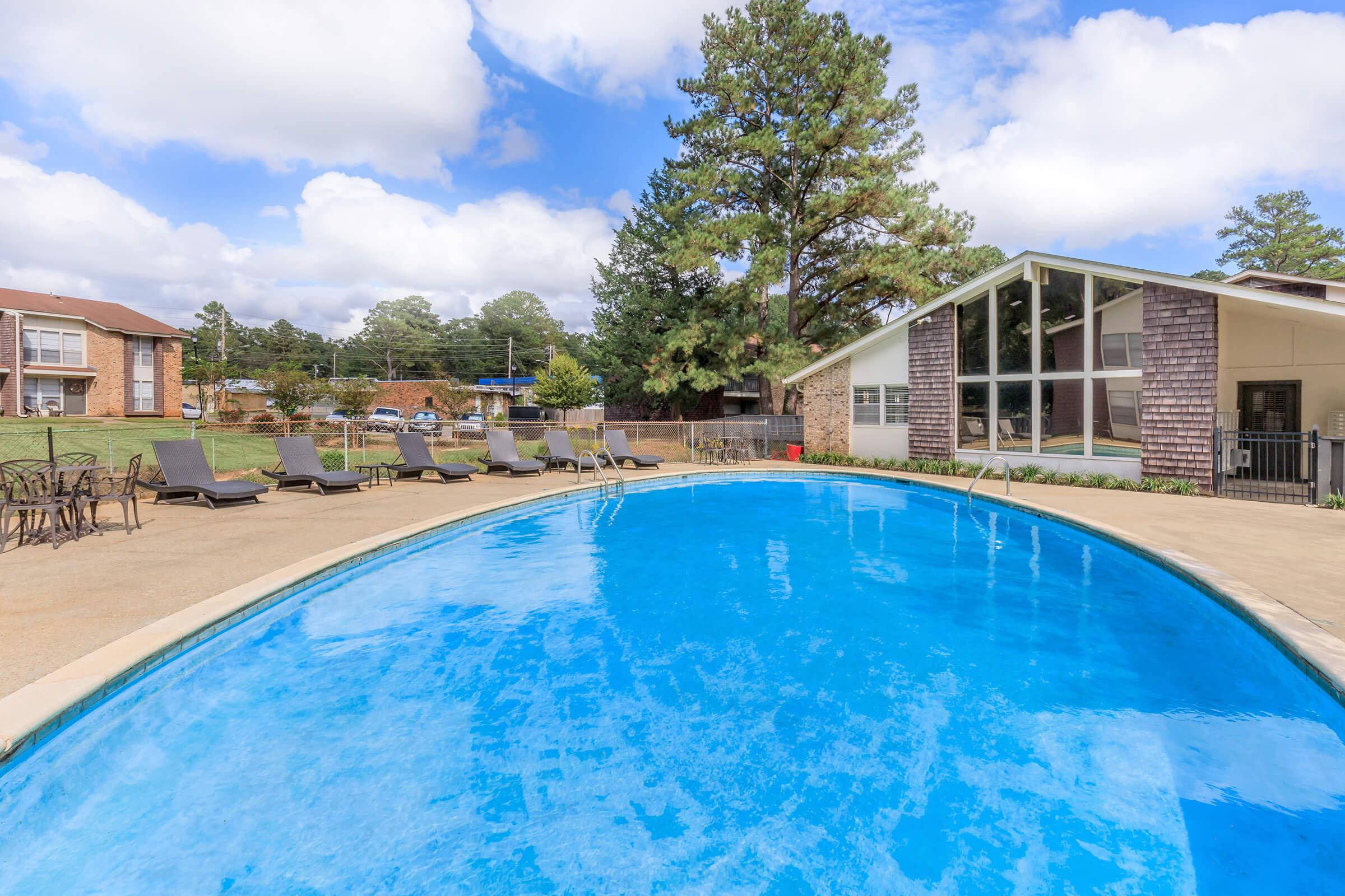 A sparkling blue swimming pool surrounded by lounge chairs, set in a residential area. In the background, a building with large windows and green trees under a partly cloudy sky. The scene is bright and inviting, perfect for relaxation and outdoor enjoyment.