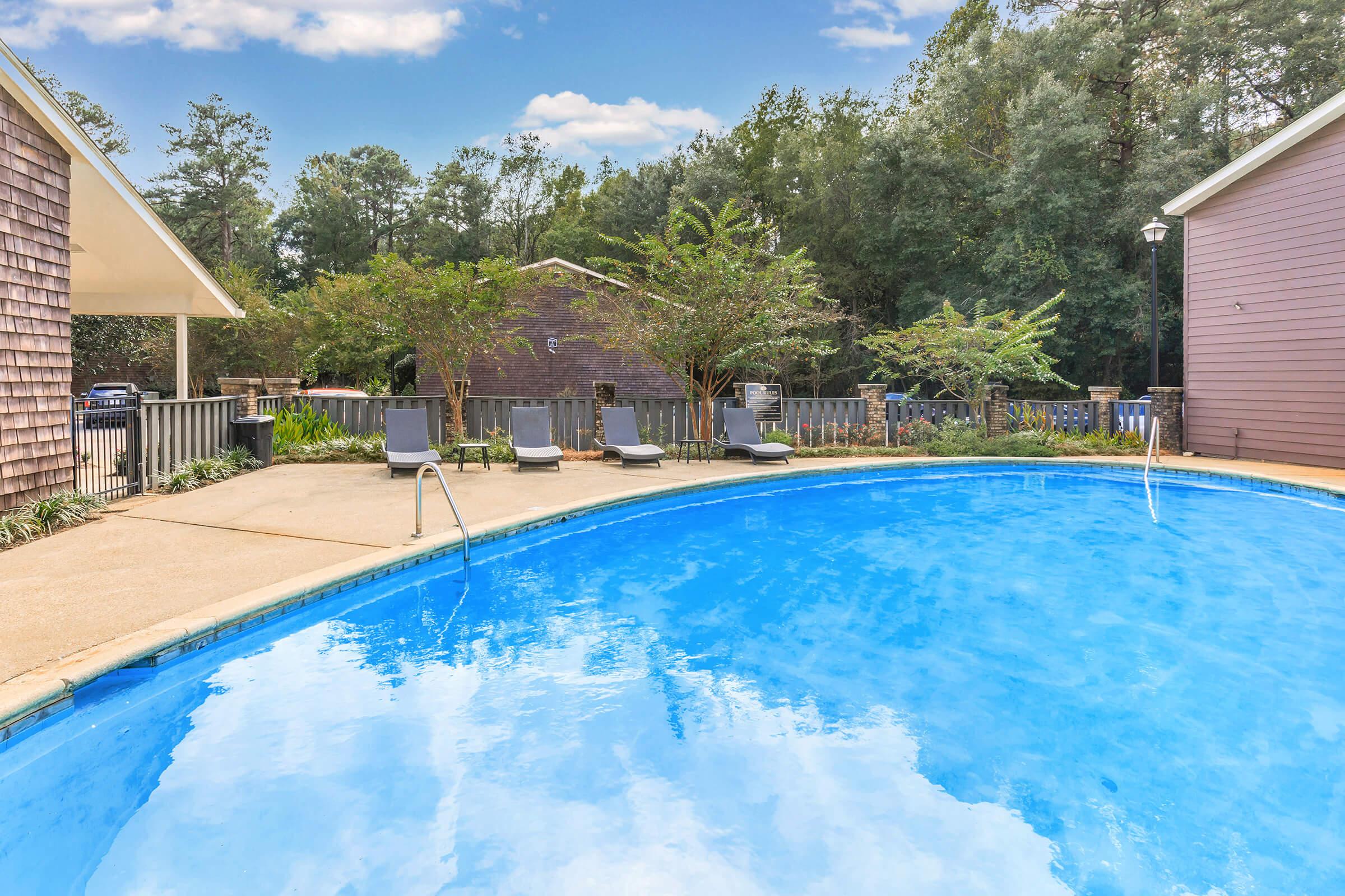 A clear blue swimming pool surrounded by lounge chairs and greenery. Several trees are visible in the background, with wooden fencing enclosing the area. The sky is partly cloudy, creating a serene outdoor atmosphere.
