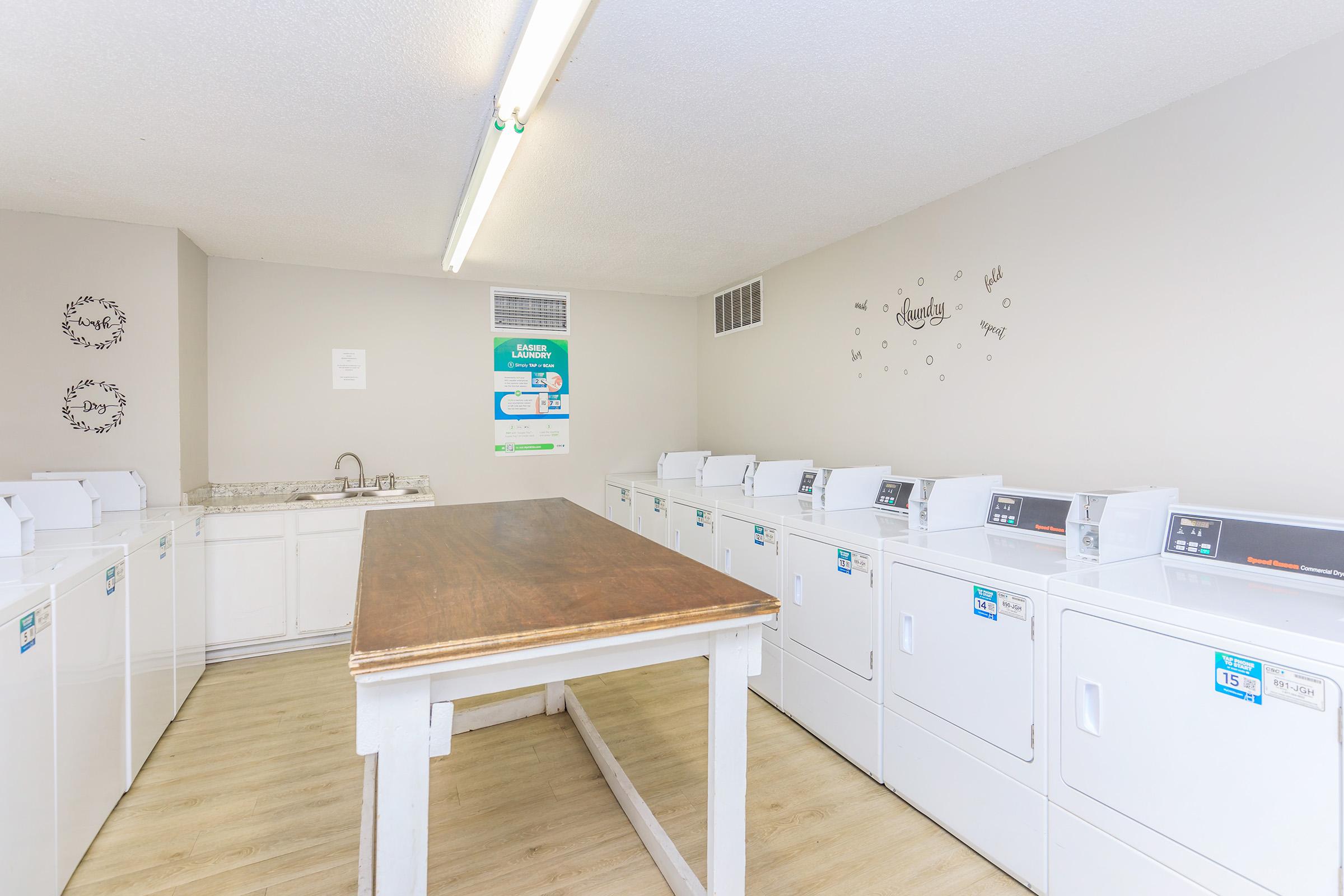 A clean and well-lit laundry room featuring several white washing machines and dryers lined against the wall. In the center, there's a wooden folding table. The walls are light-colored with decorative elements, including a motivational phrase. The space is designed for convenient laundry care.