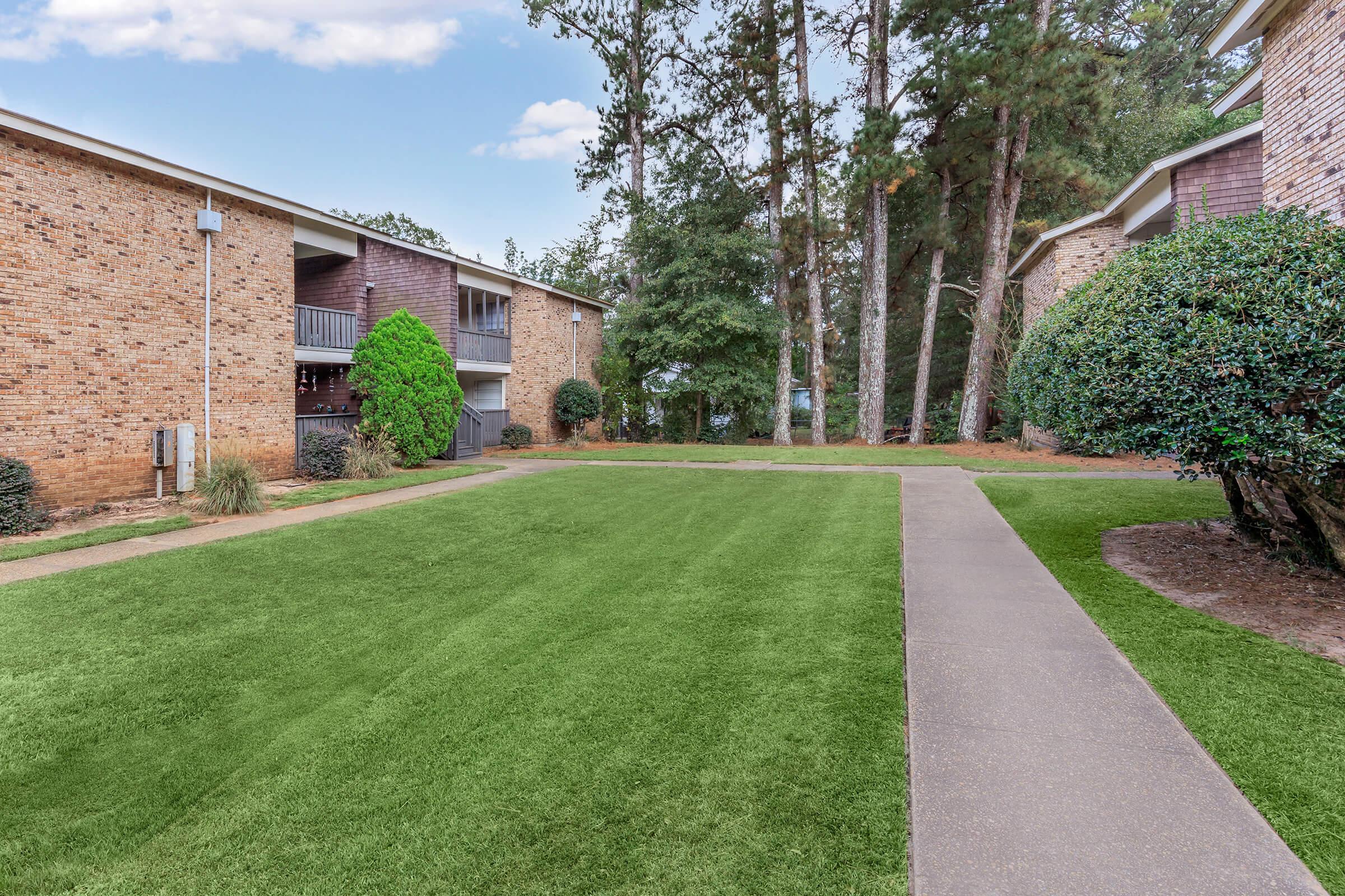 A well-maintained grassy area with a paved walkway surrounded by brick apartment buildings and trees. Lush green shrubs are present on either side of the path, creating a pleasant outdoor environment. The sky is partly cloudy, enhancing the serene atmosphere of the location.