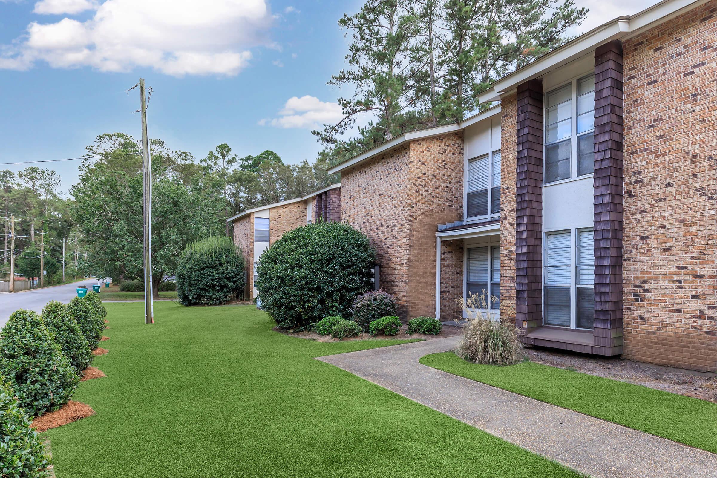 A view of a residential area featuring brick apartment buildings with large windows. The foreground includes well-maintained green grass and neatly trimmed hedges. Trees line the background, and a quiet street is visible with a utility pole. The setting appears peaceful and well-kept.