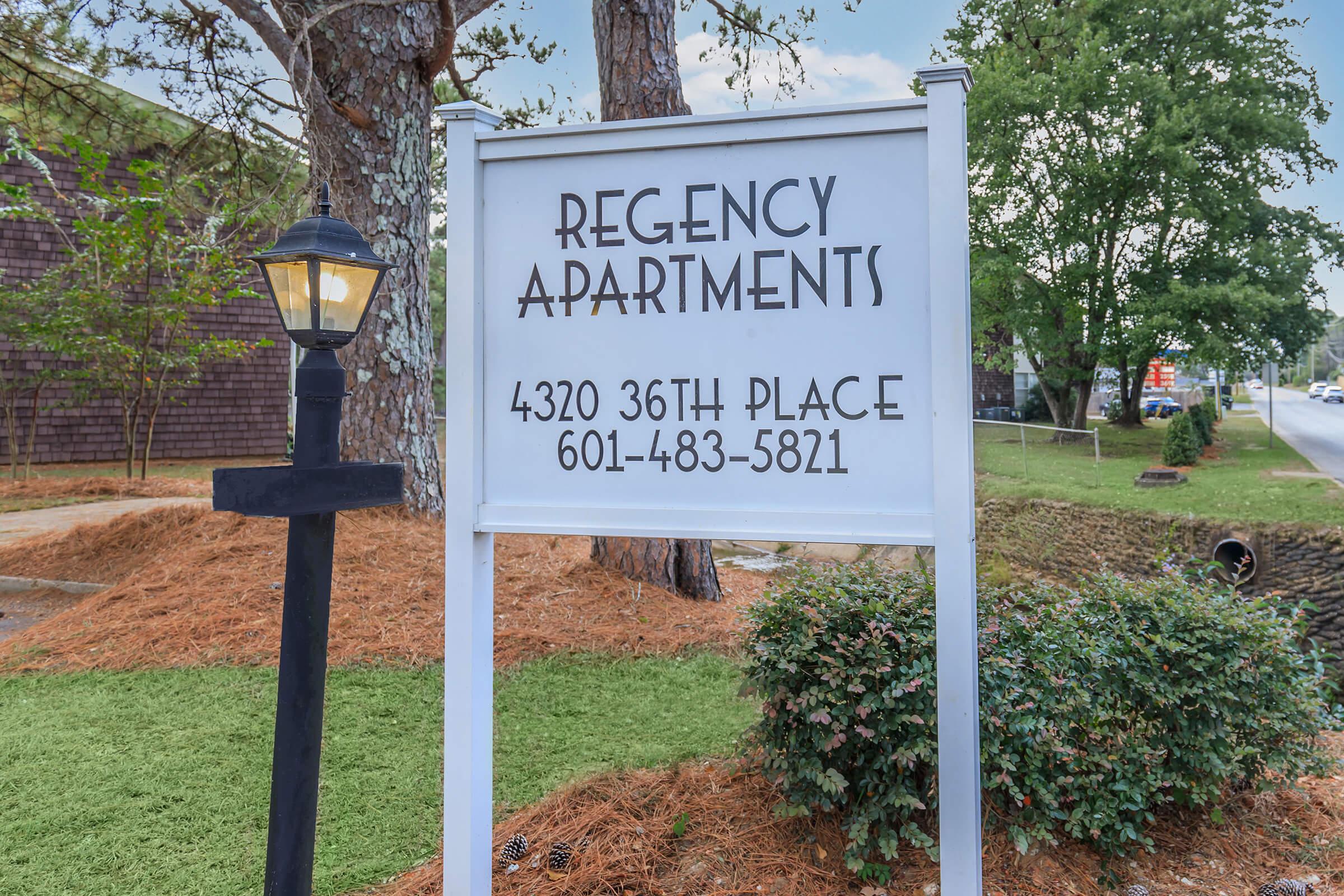 Sign for Regency Apartments displaying the address "4320 36th Place" and the phone number "601-483-5821," set among landscaped grounds with trees and a lamp post nearby. The area appears well-maintained with grass and hedges.