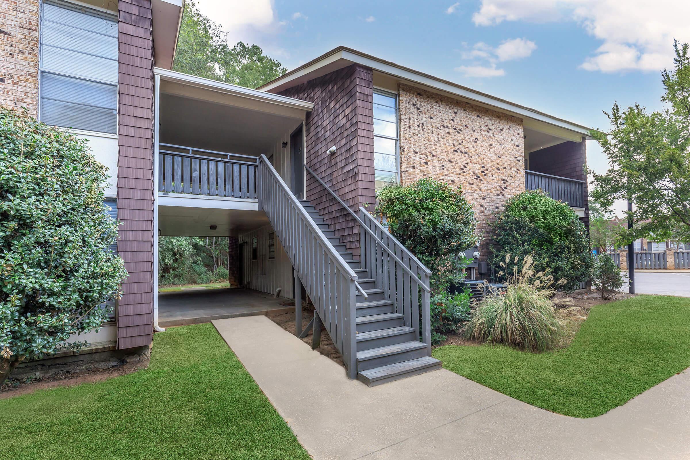 Exterior view of a two-story residential building featuring a wooden staircase leading to the second floor. The building has brick and wooden siding, with lush greenery surrounding it. A concrete pathway leads to the entrance, and there are trees and shrubs landscaping the area.