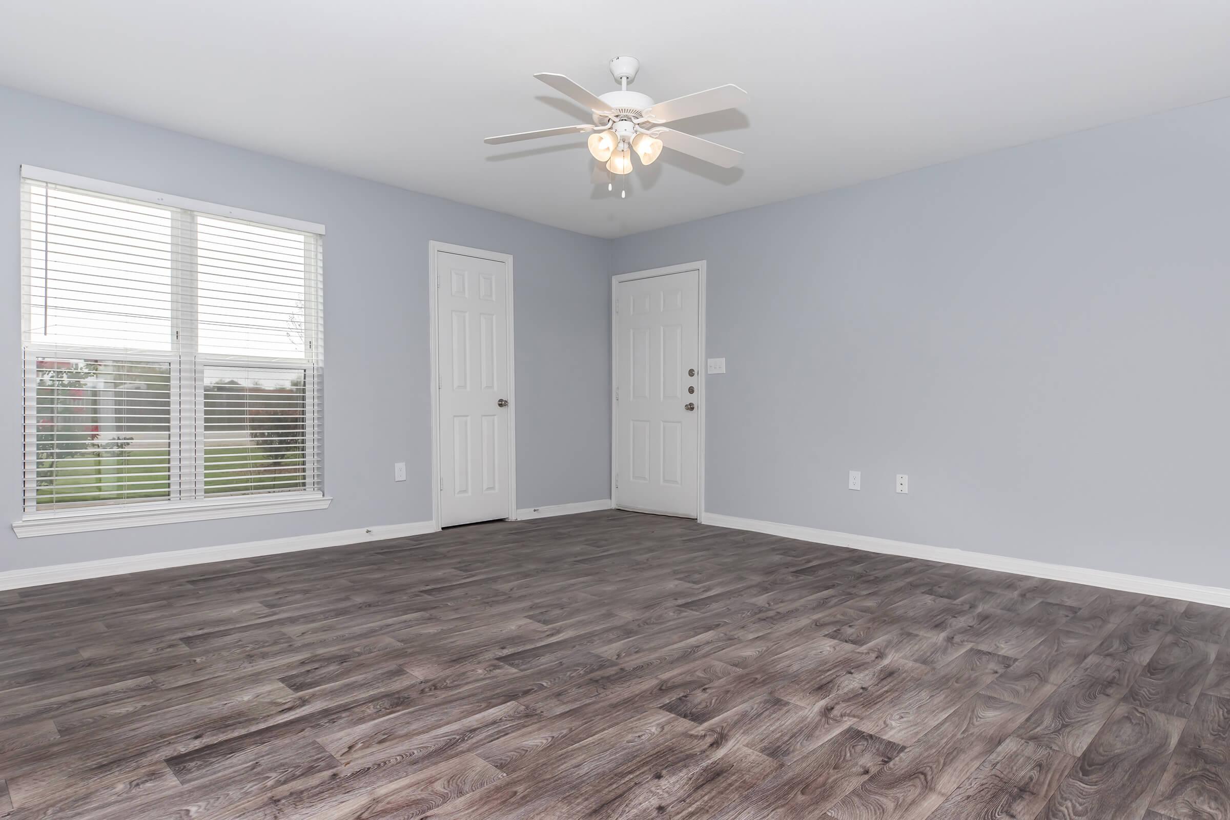 A vacant room with light gray walls and a ceiling fan. It has two closed doors, one on the right and one on the left, and a large window with blinds allowing natural light. The floor is a wood-like laminate in a dark hue. The overall atmosphere is bright and spacious.