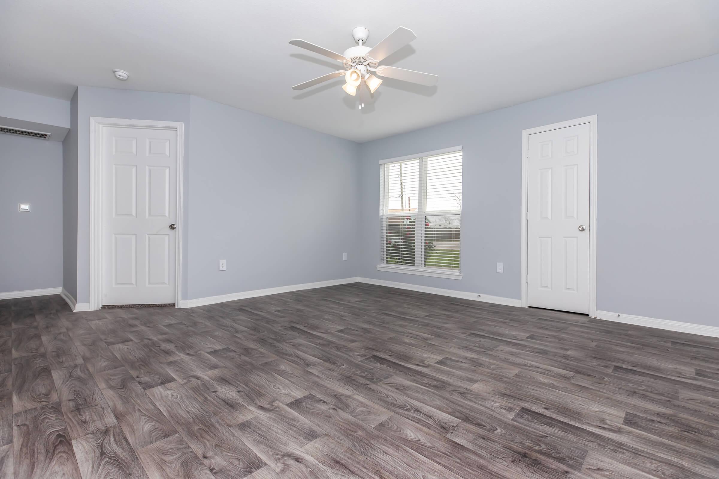 Spacious room with light gray walls and a ceiling fan. Features two closed white doors and a window with blinds, allowing natural light. The floor is covered in dark wood laminate, creating a warm and inviting atmosphere.