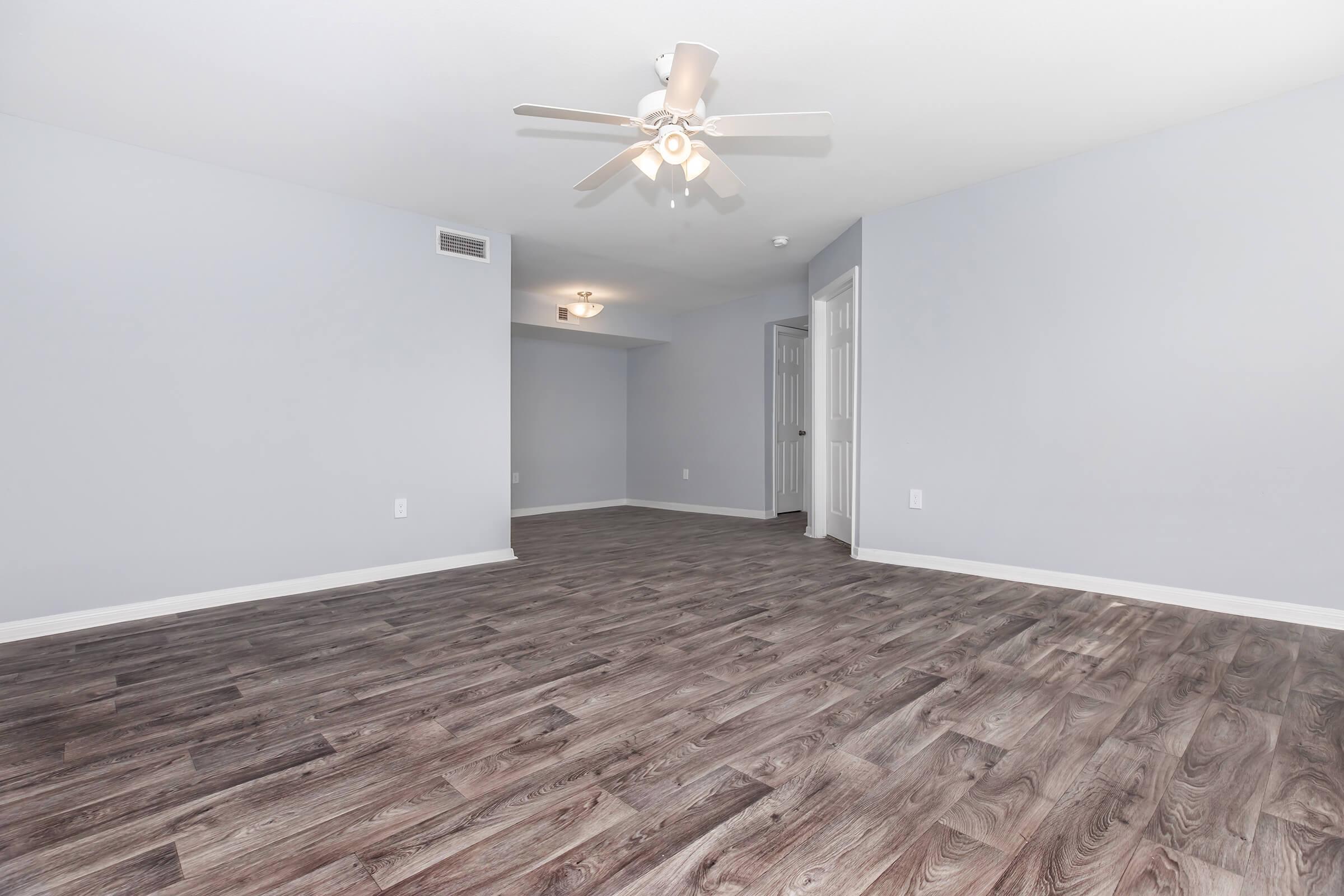 A spacious, empty room with light gray walls and a wooden floor. A ceiling fan is visible. The room has a doorway leading to another space, providing a bright and airy atmosphere. There are no furnishings, making it a blank canvas for customization.