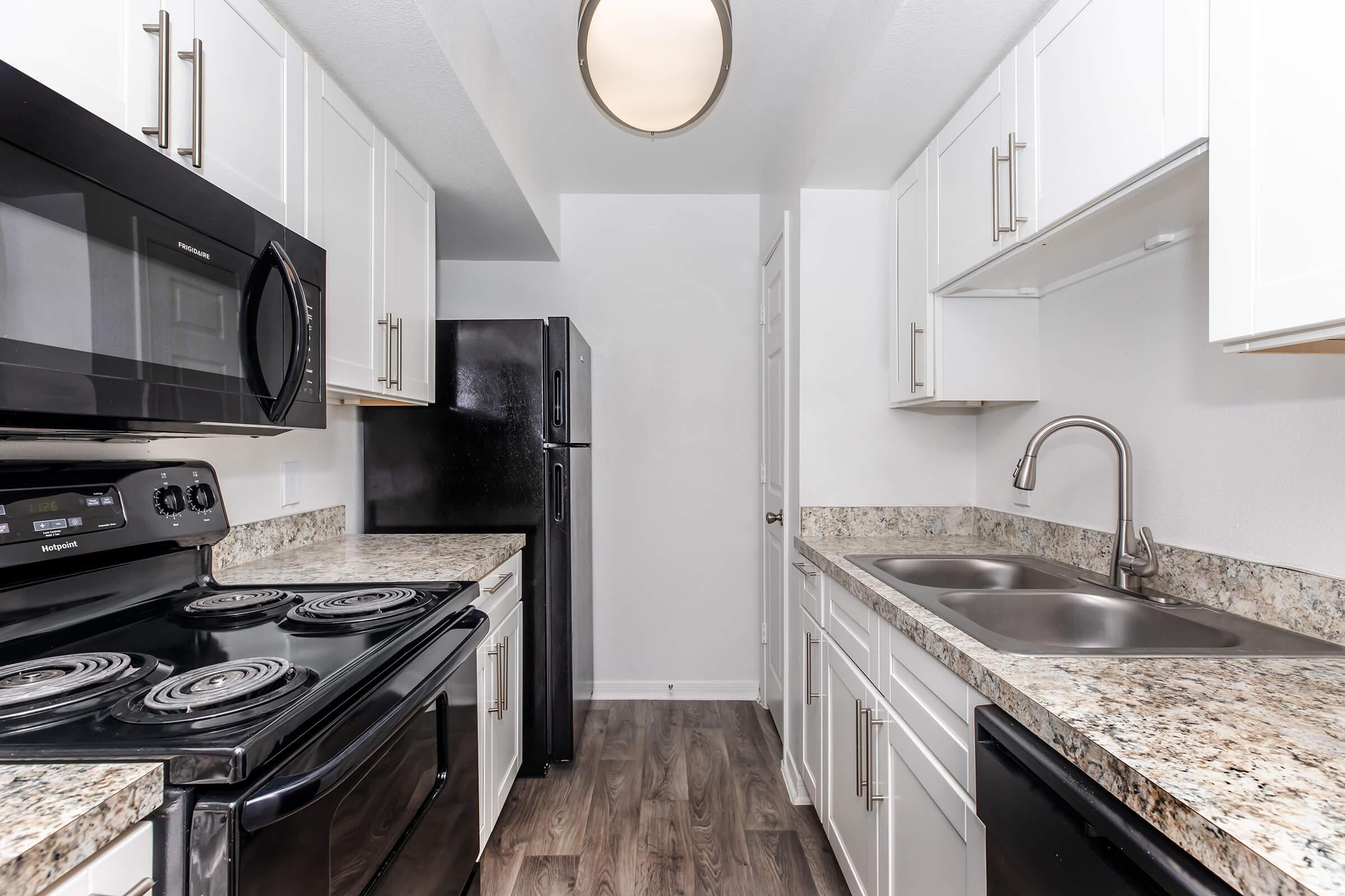 A modern kitchen featuring stainless steel appliances, including a microwave, stove, and black refrigerator. The countertops are made of speckled granite, and there are white cabinetry and a double sink. The space is well-lit with a ceiling fixture and has a contemporary aesthetic.