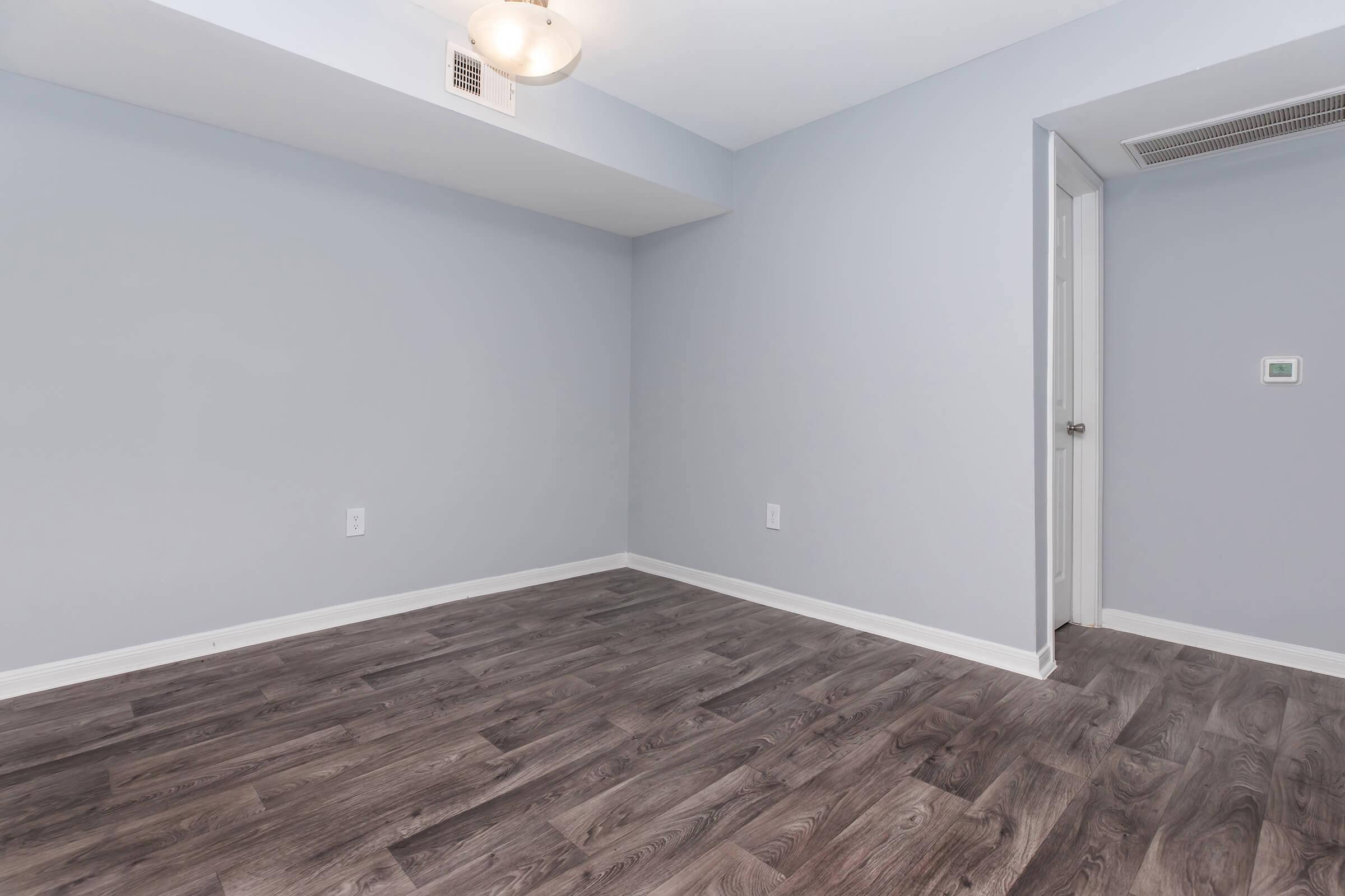 Empty room with light gray walls and a wooden floor. A ceiling light fixture is visible. There’s a door on the right leading to another space and a small wall panel near the door. The overall atmosphere is clean and minimalistic, suggesting a fresh living space ready for furnishings.