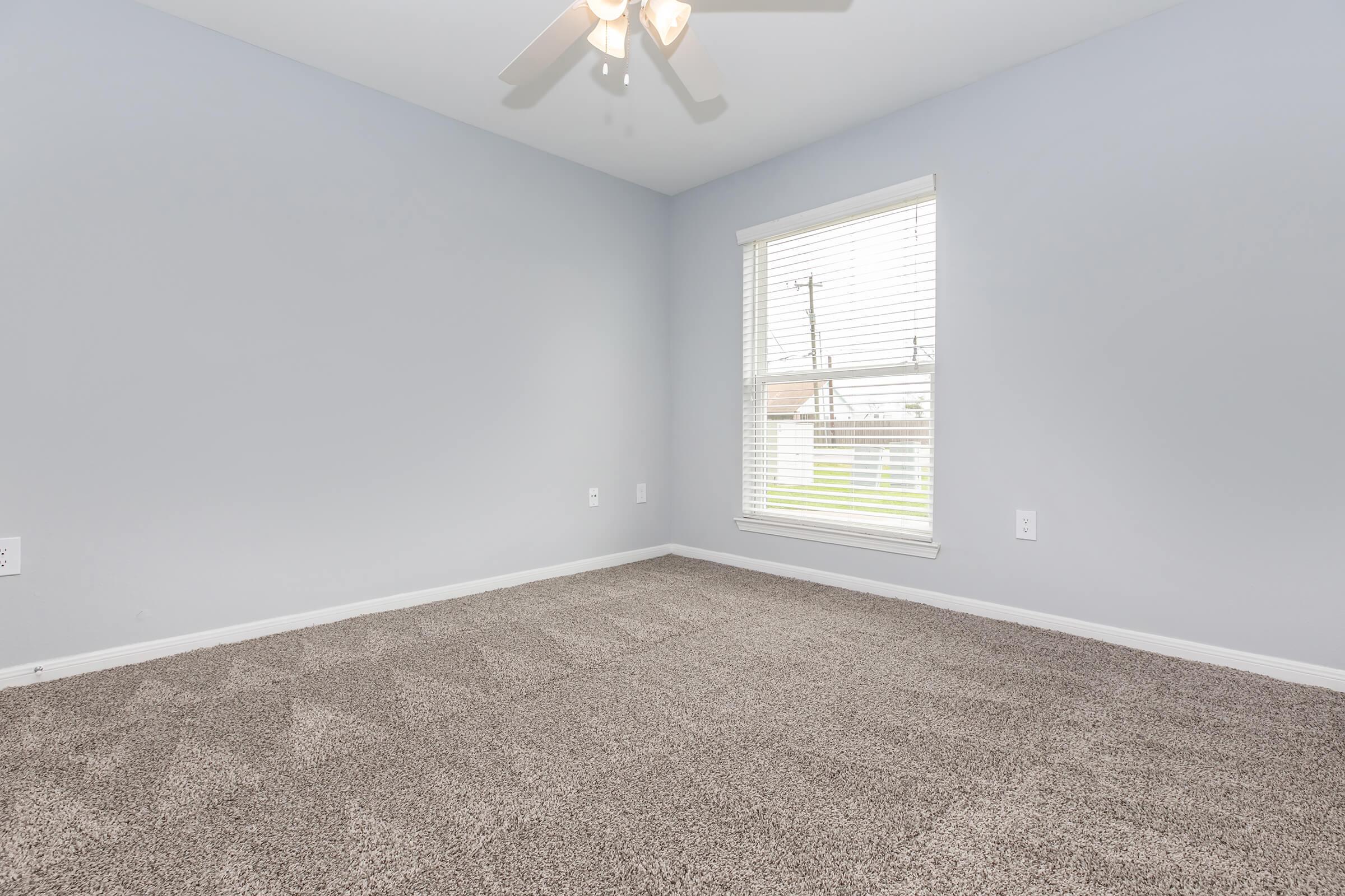 Empty room with light gray walls and a ceiling fan. The floor is covered in plush, beige carpet. A window with white blinds lets in natural light, illuminating the space. No furniture or decorations are present.