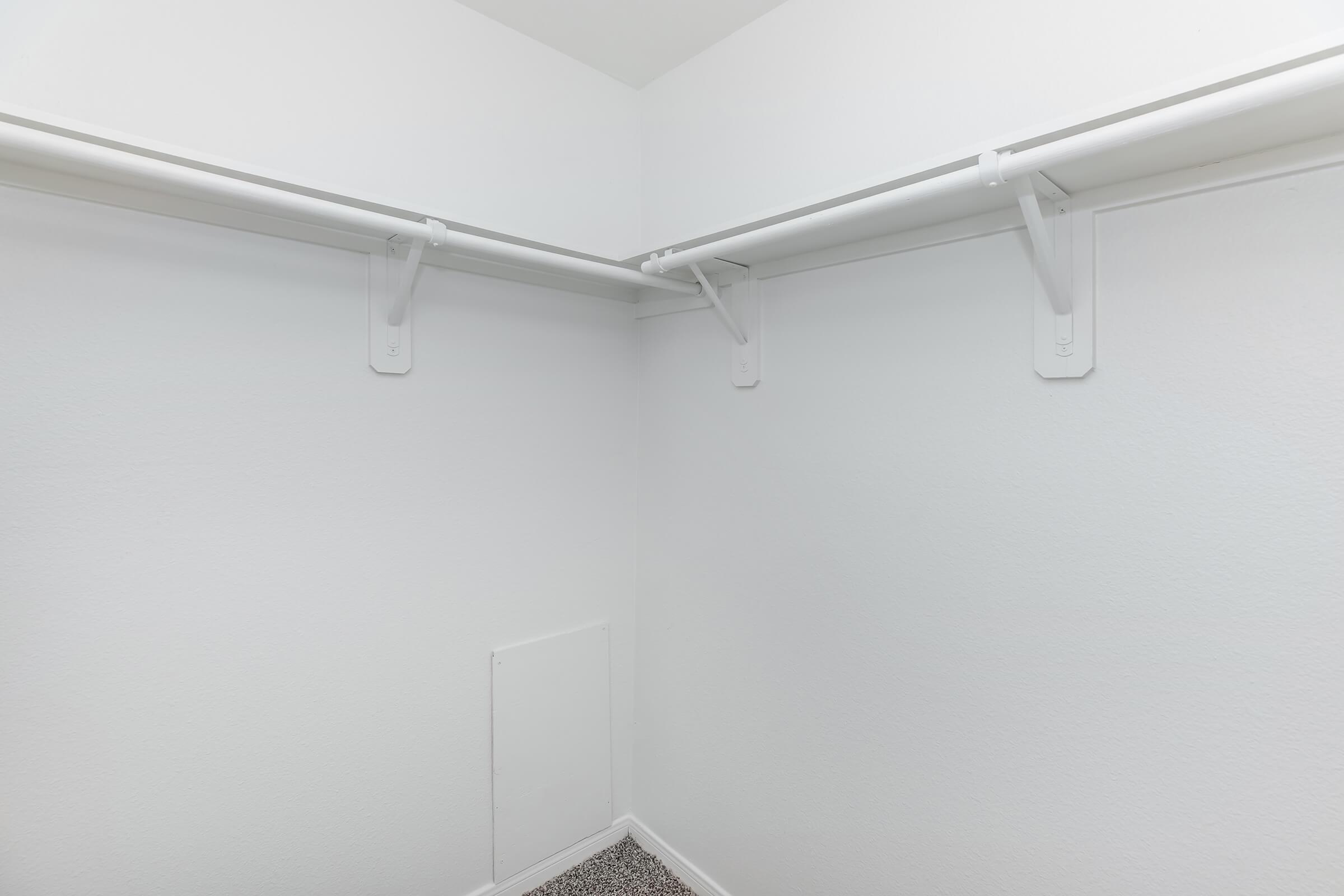 A small, empty closet with two white shelving rods mounted on the walls. The walls are plain white, and the floor is covered with small, dark-colored tiles. There is a small access panel on one wall, adding to the minimalistic design of the space.