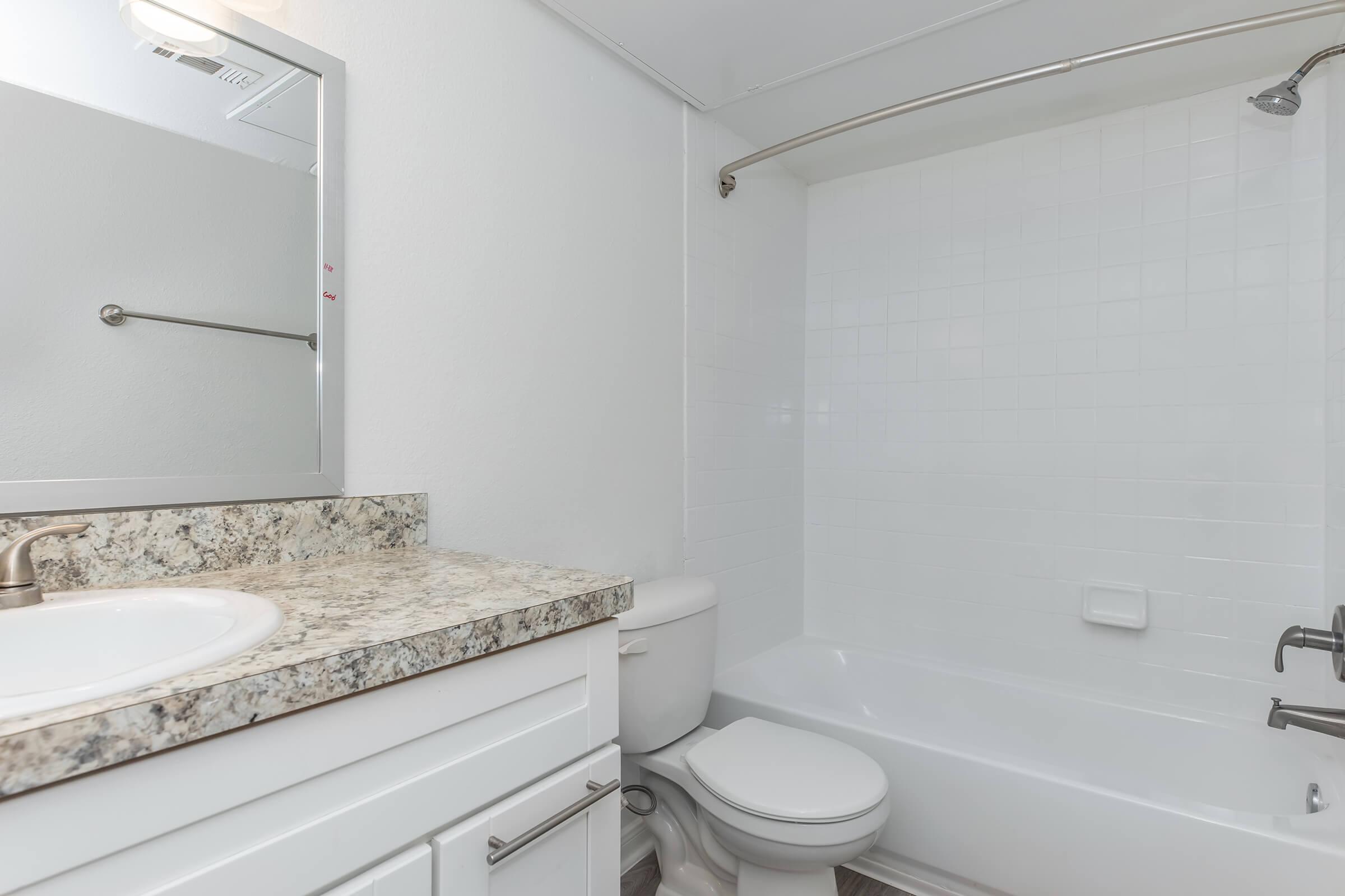 A modern bathroom featuring a white bathtub and shower combination, a white toilet, and a granite countertop sink with a mirror above it. The walls are painted white, and the space is well-lit, providing a clean and fresh look.