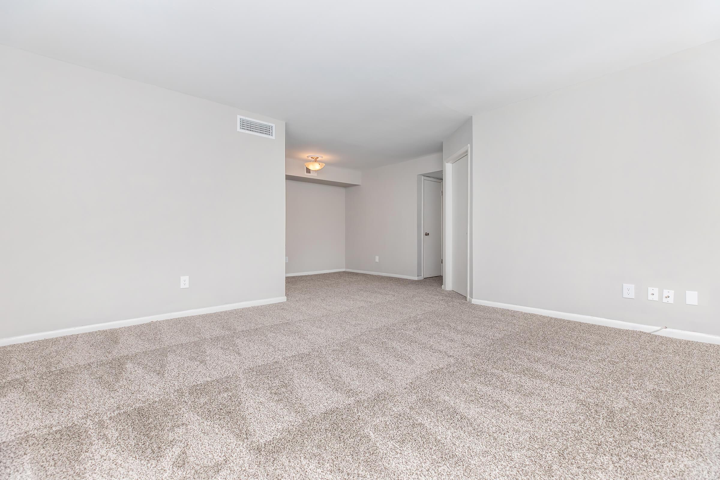 A spacious, light-colored room with beige carpet flooring. The walls are painted in a neutral tone, and there is a doorway leading to another area. A ceiling light fixture provides illumination, and there are no furnishings in the space, giving it an open, airy feel.