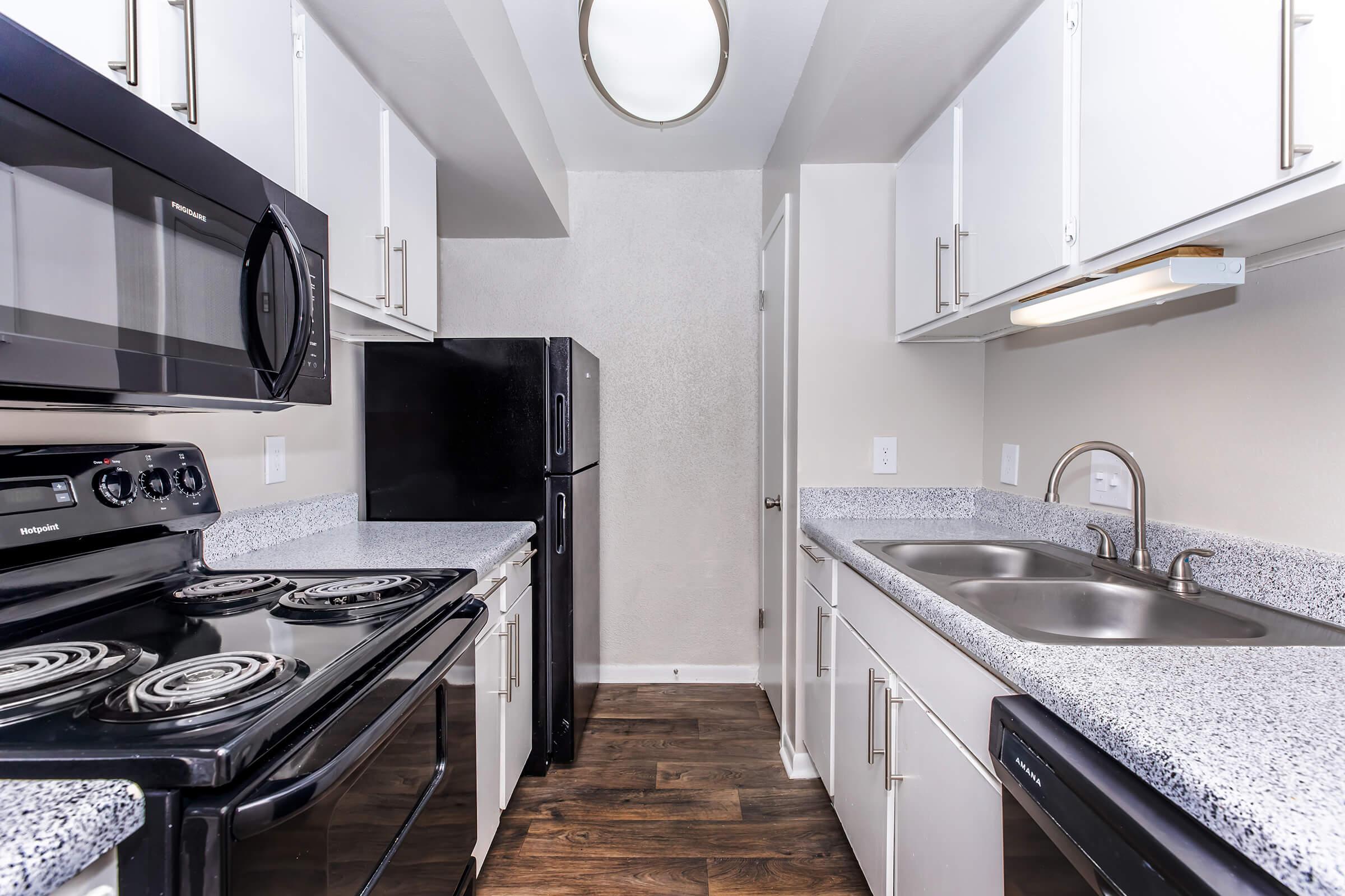 A modern kitchen featuring a black stove, microwave, and refrigerator, with white cabinets and a gray countertop. The space also includes a double sink and a light fixture, set against a neutral wall color. The flooring is dark wood.