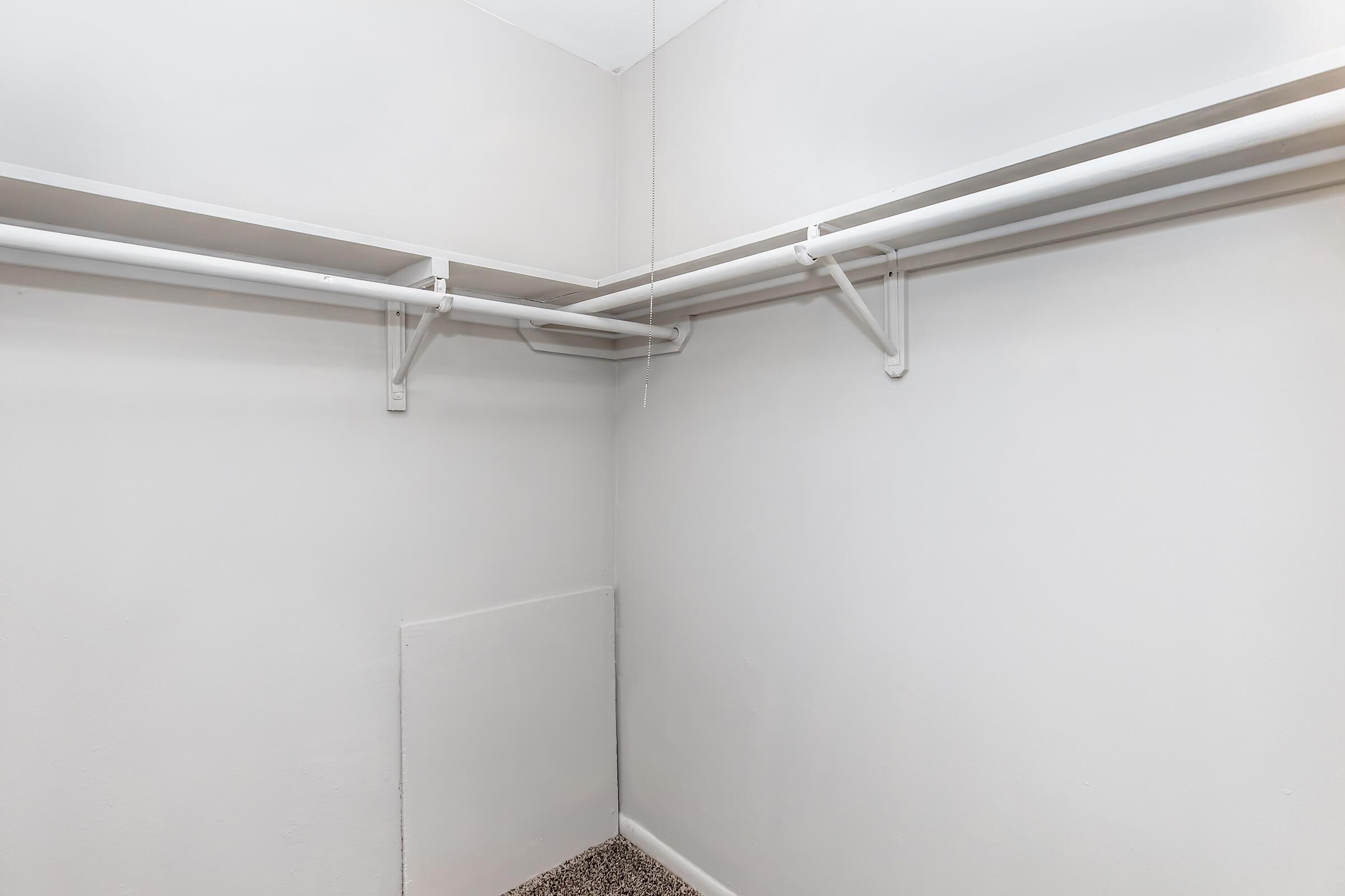 Empty closet with white walls and two horizontal hanging rods. The floor is covered in beige carpet, and there is a small panel on one wall. The space is well-lit and features a minimalistic design.