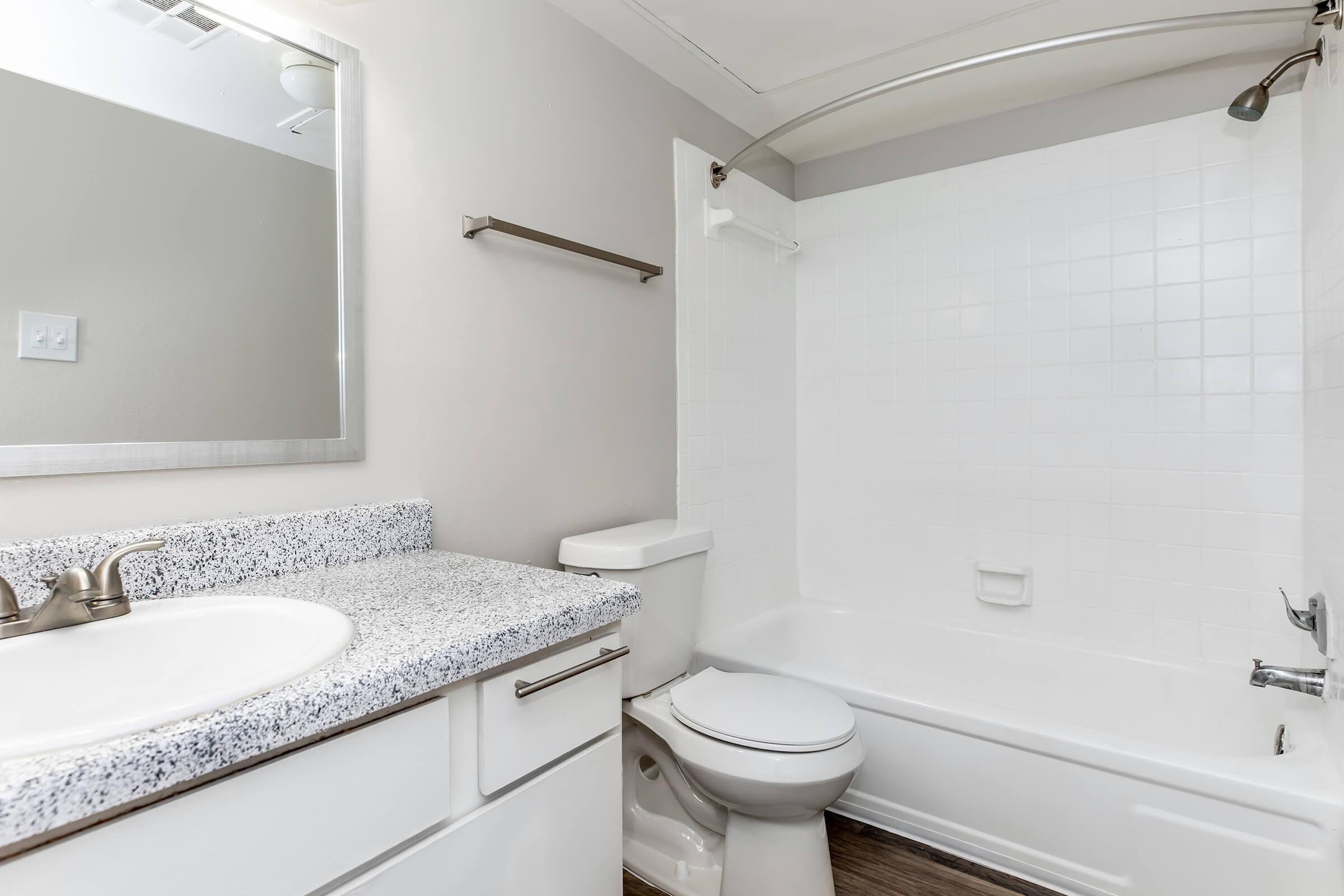 A clean, modern bathroom featuring a white bathtub with an adjacent shower, light gray walls, a marble countertop sink with a mirror above, and a toilet. The space has a neutral color palette and includes a towel rack, creating a bright and airy atmosphere.