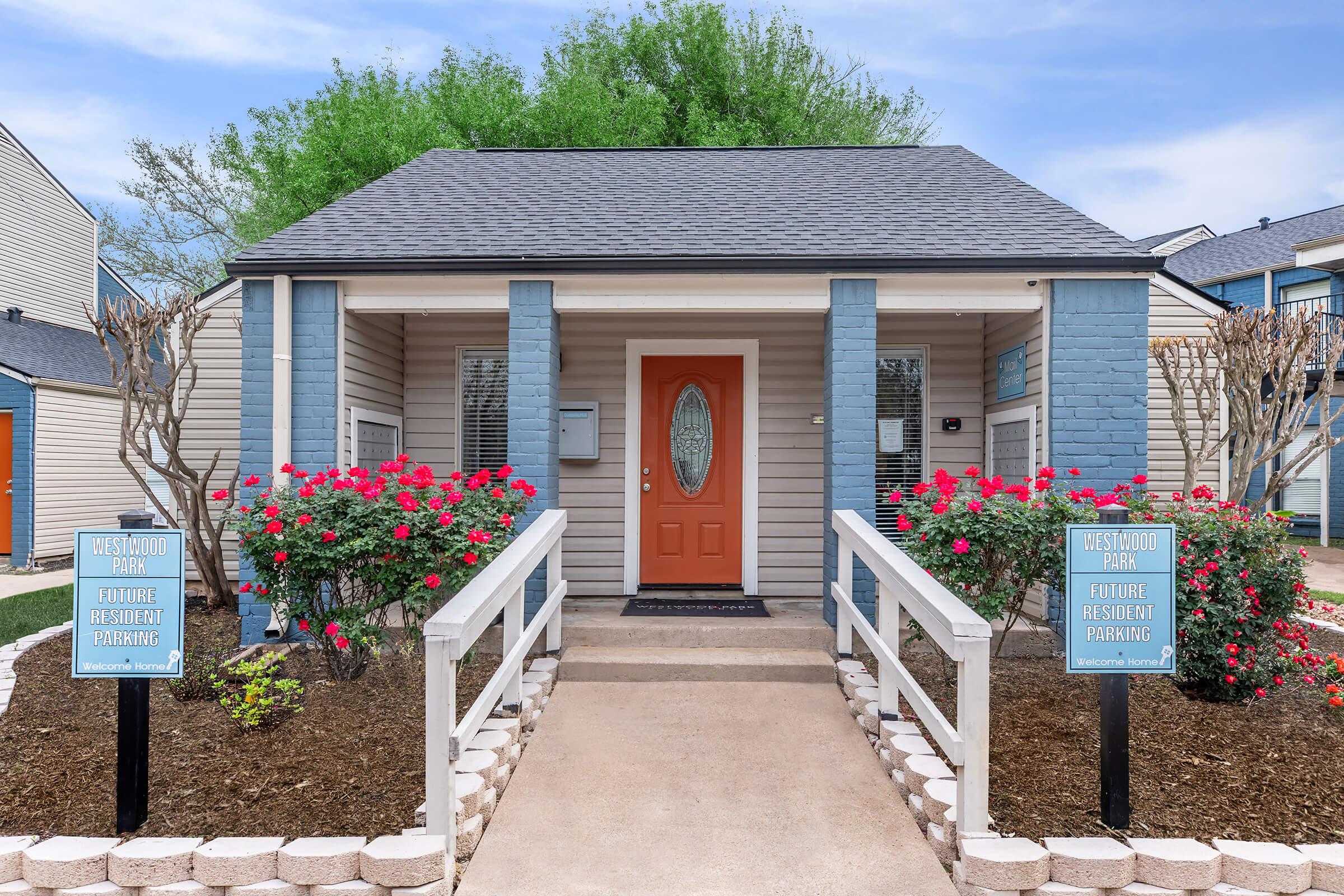 A small house with a welcoming front porch, featuring an orange front door. The entrance is surrounded by blooming rose bushes and a pathway lined with decorative stones. Two signs are positioned on either side of the path, indicating parking information for residents. Lush green trees are visible in the background.