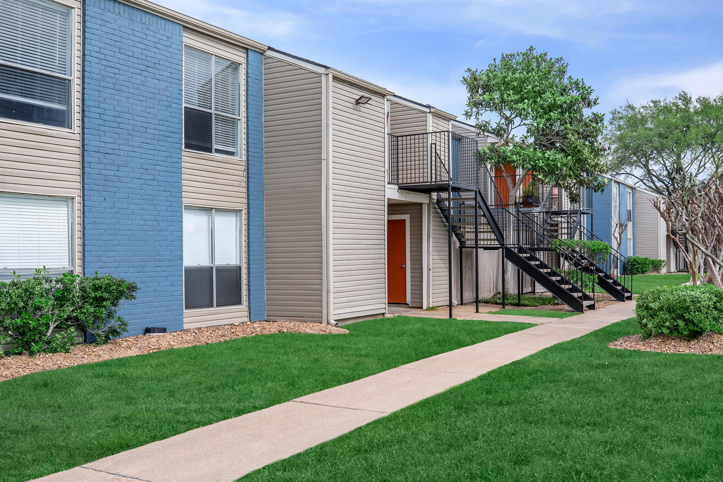 A photo of a modern apartment complex featuring two-story buildings with a mix of blue and gray siding. There is a pathway leading to a staircase that connects the units. Lush green grass and neatly trimmed shrubs surround the buildings, creating a pleasant outdoor environment.
