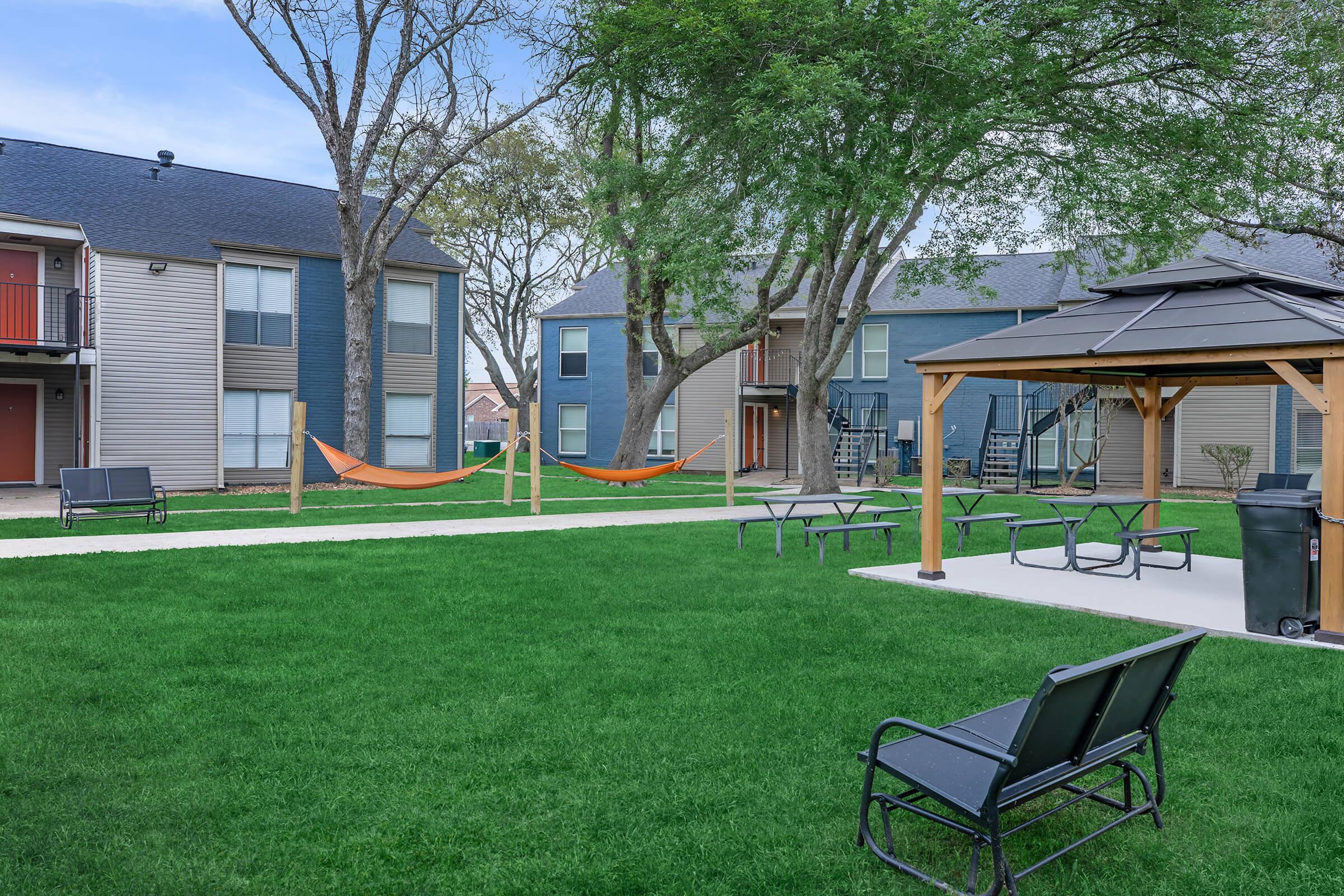 A well-maintained outdoor courtyard featuring green grass, two hammocks strung between trees, and several seating areas with picnic tables and benches. A gazebo is also present, along with a grill, creating a welcoming space for relaxation and social gatherings.