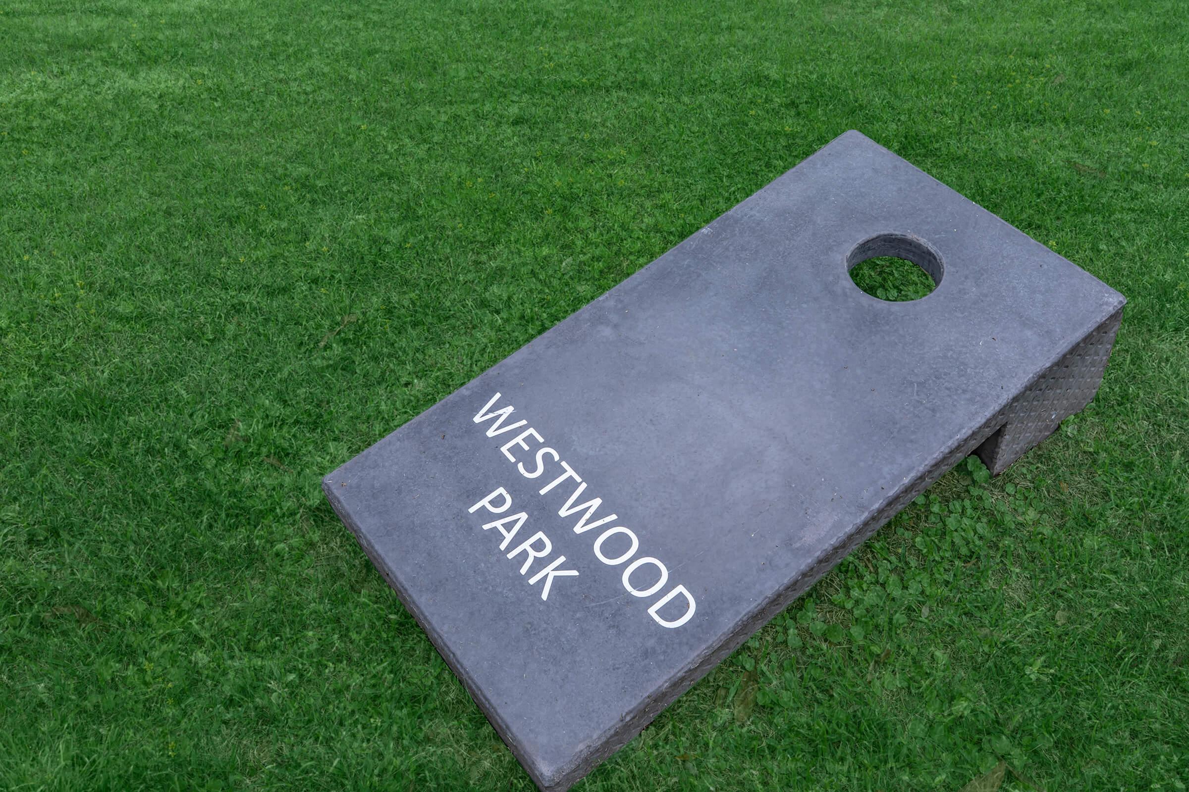 A gray cornhole board resting on green grass, with "WESTWOOD PARK" displayed prominently on the surface.