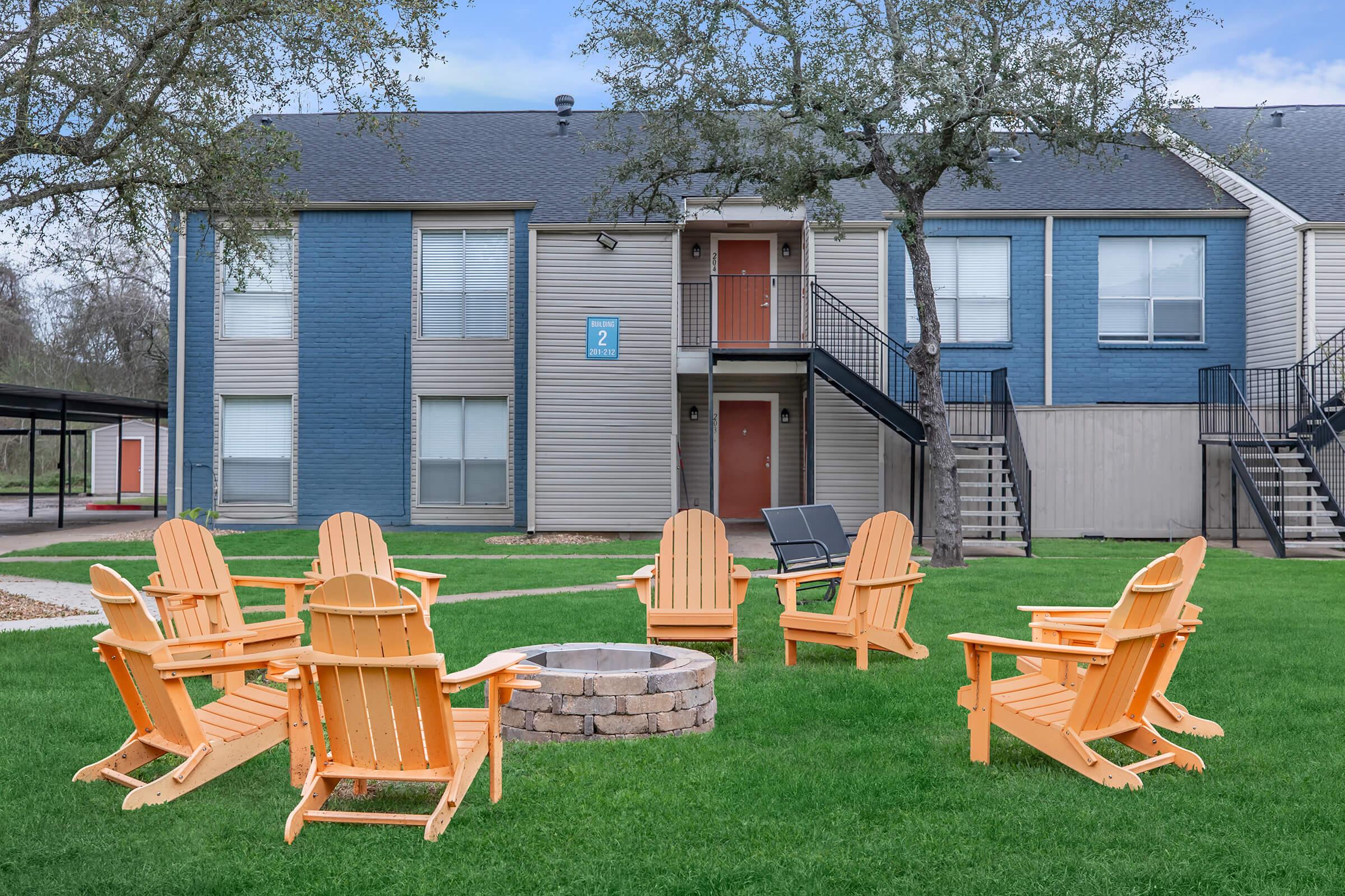 A cozy outdoor seating area with several orange Adirondack chairs arranged around a stone fire pit. In the background, a two-story apartment building features a mix of blue and beige siding, with a staircase leading to the upper level and a door marked with the number 2. Green grass surrounds the seating area.