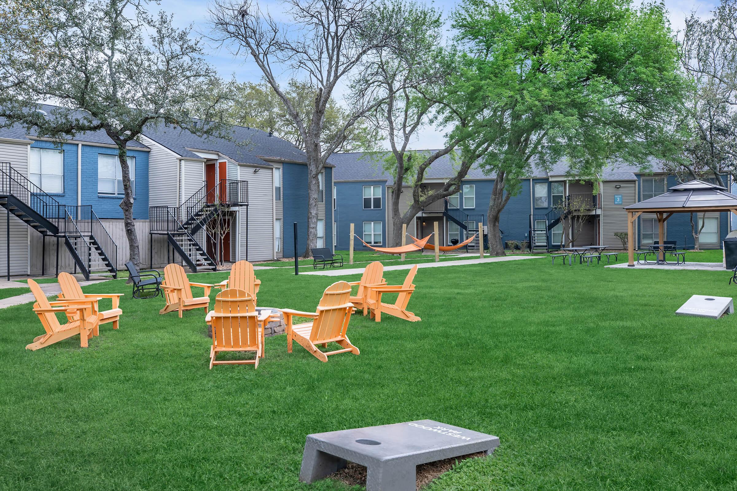 A well-maintained green courtyard featuring several orange Adirondack chairs arranged around a fire pit, surrounded by trees and two apartment buildings. In the background, there is a hammock hanging between trees and a gazebo, creating a relaxing outdoor space for residents.