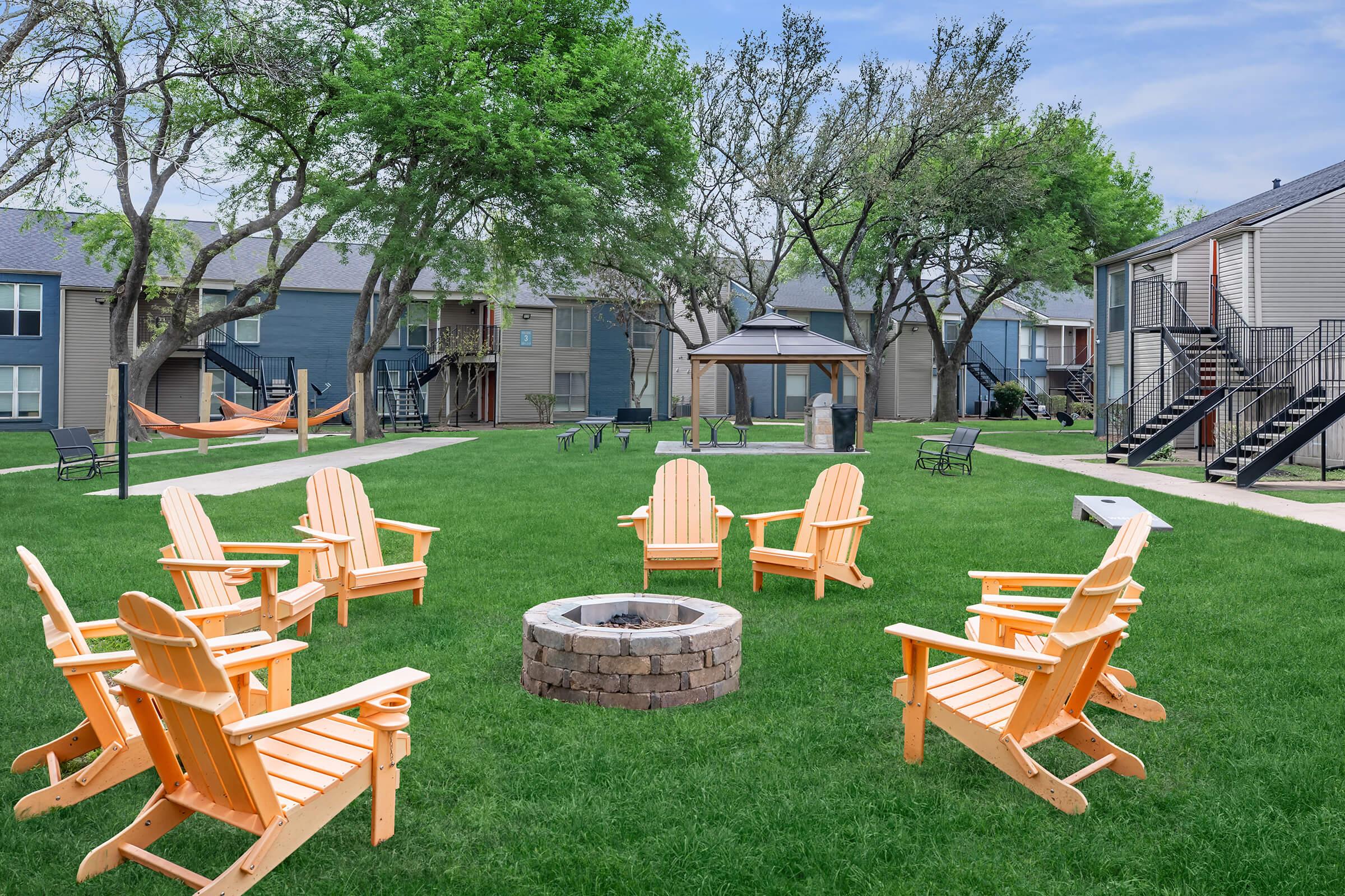 A circular arrangement of bright orange Adirondack chairs around a stone fire pit, set on a lush green lawn. In the background, there are residential buildings with balconies, a hammock between two trees, and additional seating throughout the area, under a clear blue sky.