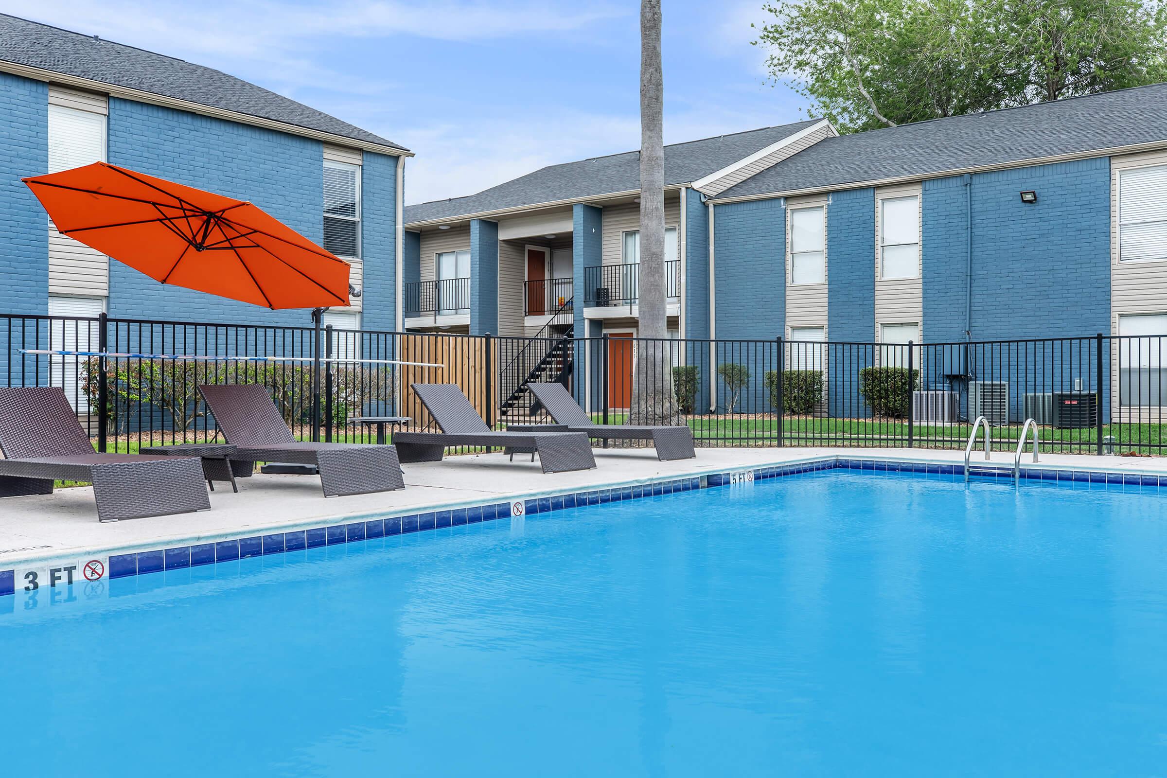 A swimming pool with clear blue water, surrounded by lounge chairs and an orange umbrella. In the background, there are two blue apartment buildings with multiple stories and a well-maintained lawn. The scene is bright and inviting, suggesting a relaxing outdoor area.