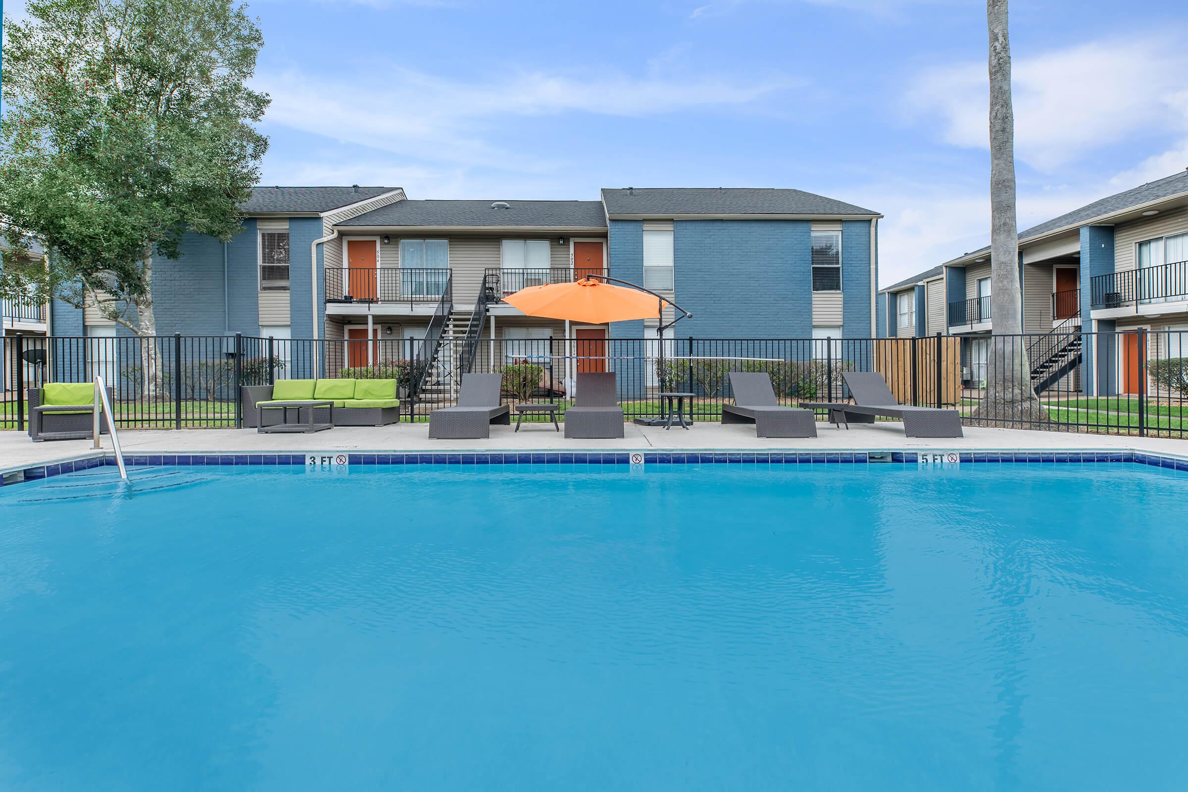 A clear blue swimming pool in the foreground with lounge chairs and an orange umbrella. In the background, a multi-story apartment building features balconies and well-maintained landscaping, against a clear blue sky.