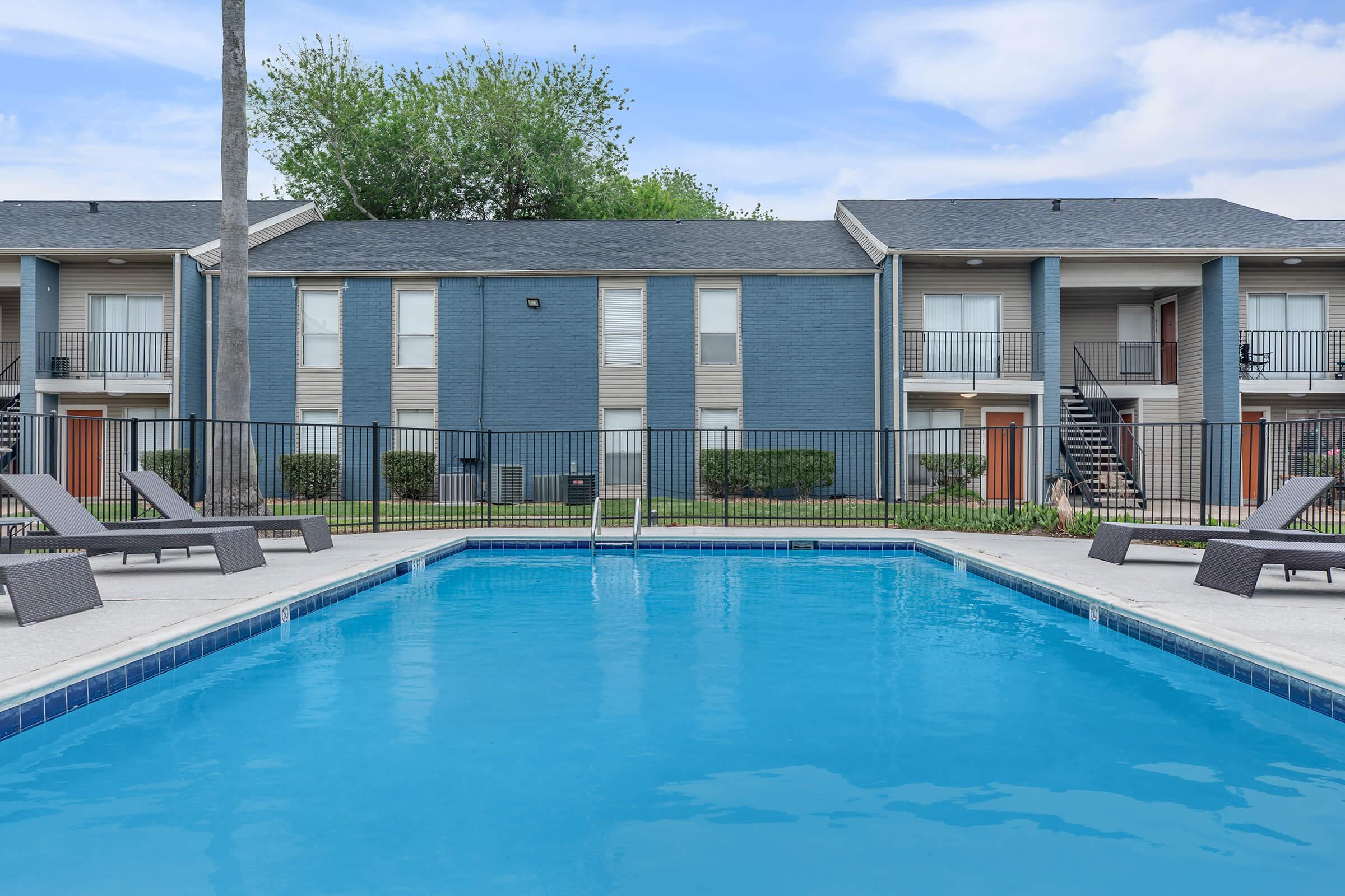 A clear swimming pool surrounded by lounge chairs and a black iron fence, with a two-story apartment building in the background featuring a gray exterior and balconies. Lush green trees are visible above the building against a bright blue sky.
