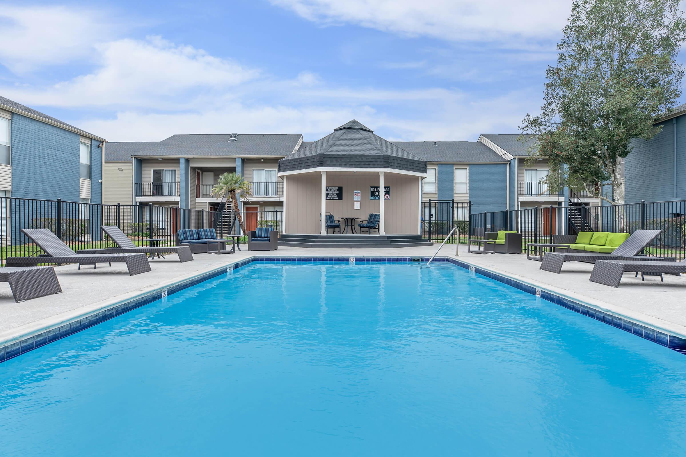 A clear blue swimming pool surrounded by lounge chairs, with a gazebo structure in the background. The area is part of an apartment complex featuring several buildings. Lush trees are located nearby, and the sky is partly cloudy.