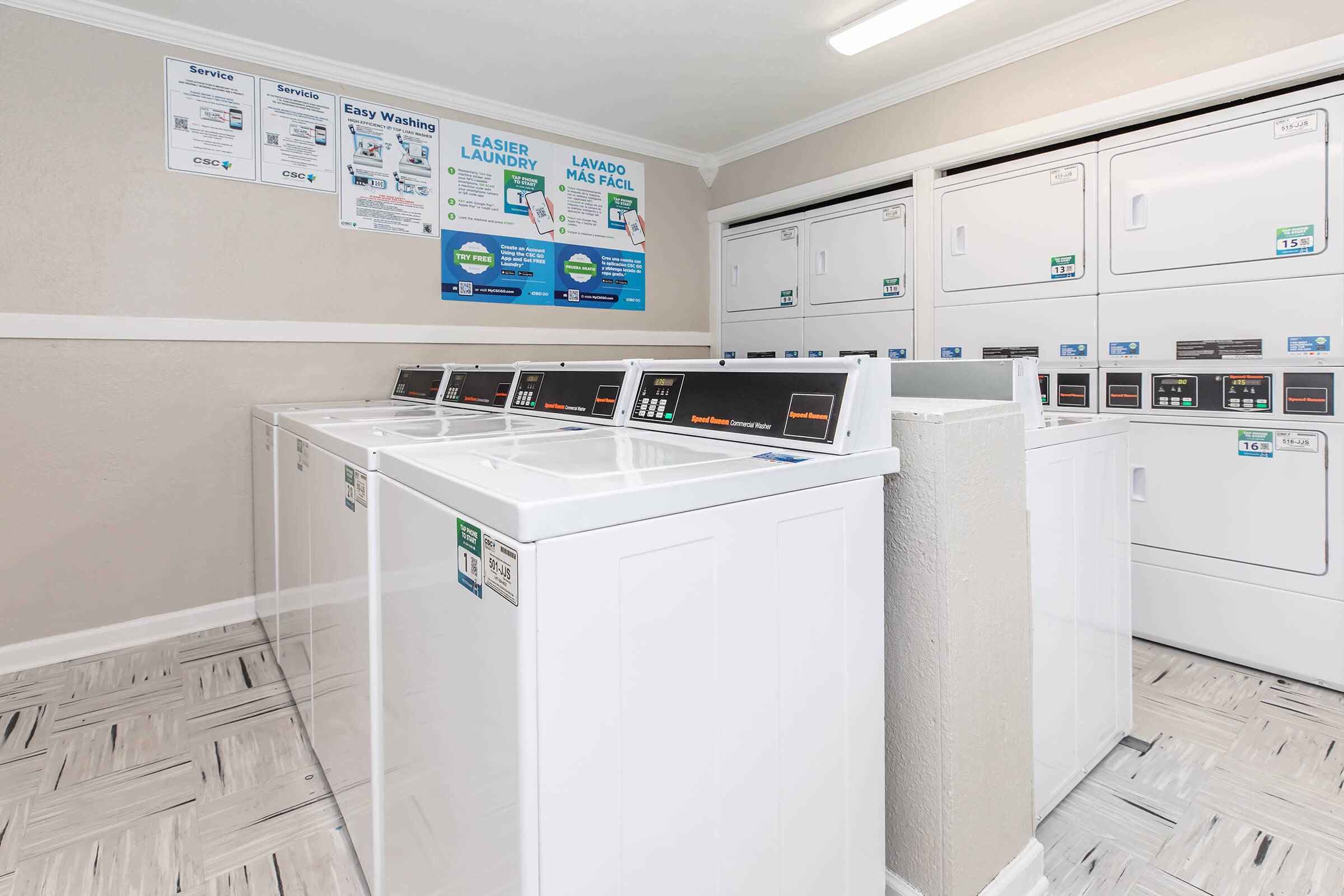 A clean, well-lit laundromat featuring several white washing machines and dryers lined up against a wall. Informational signage about services and laundry tips is visible on the wall, with a neutral color scheme and tiled flooring, creating a welcoming atmosphere for customers.