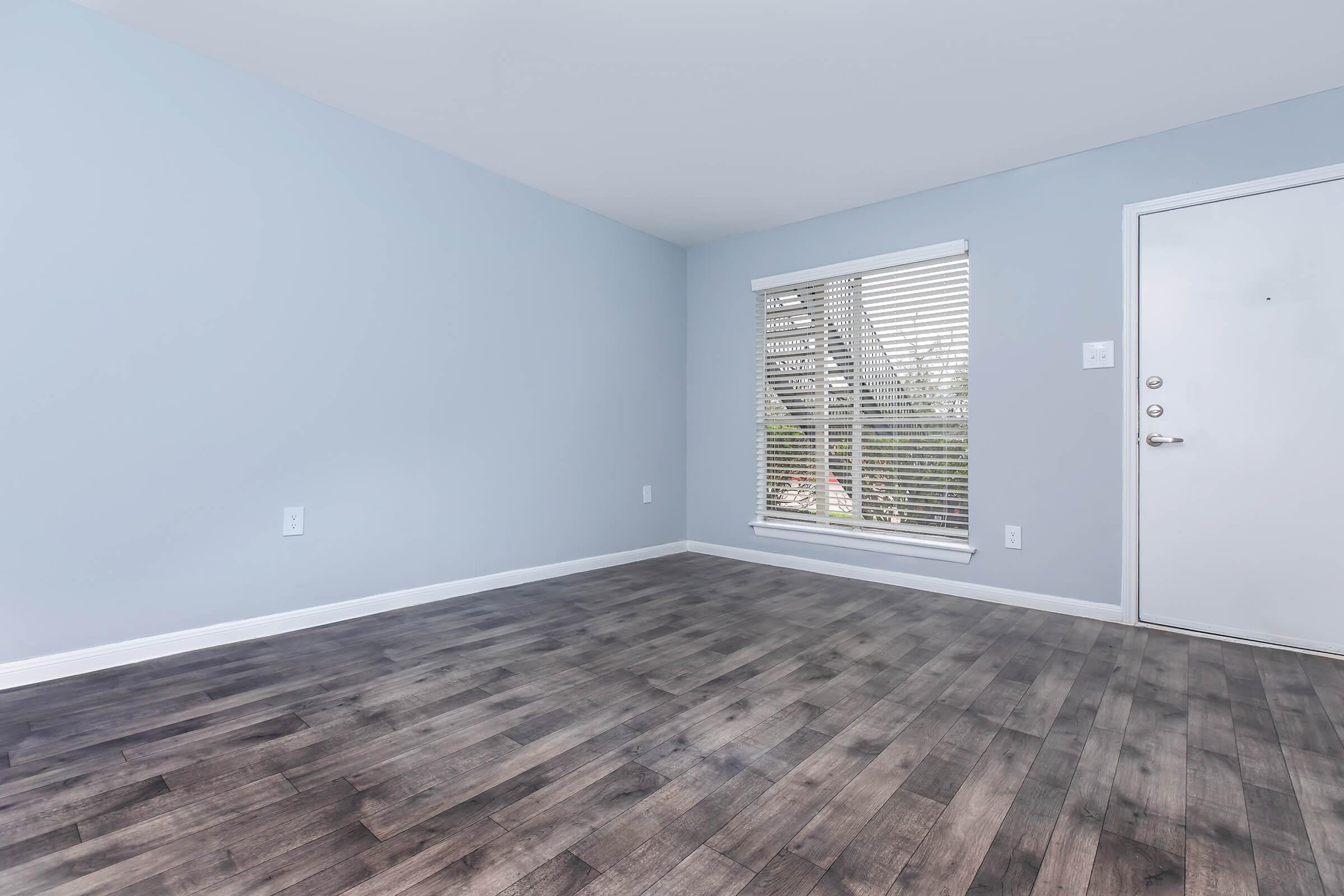 A bright, empty room with light blue walls and a modern, dark wood-like floor. There's a window covered with white blinds allowing natural light to enter, and a plain white door visible on the right side. The space is clean and uncluttered, creating a minimalist atmosphere.