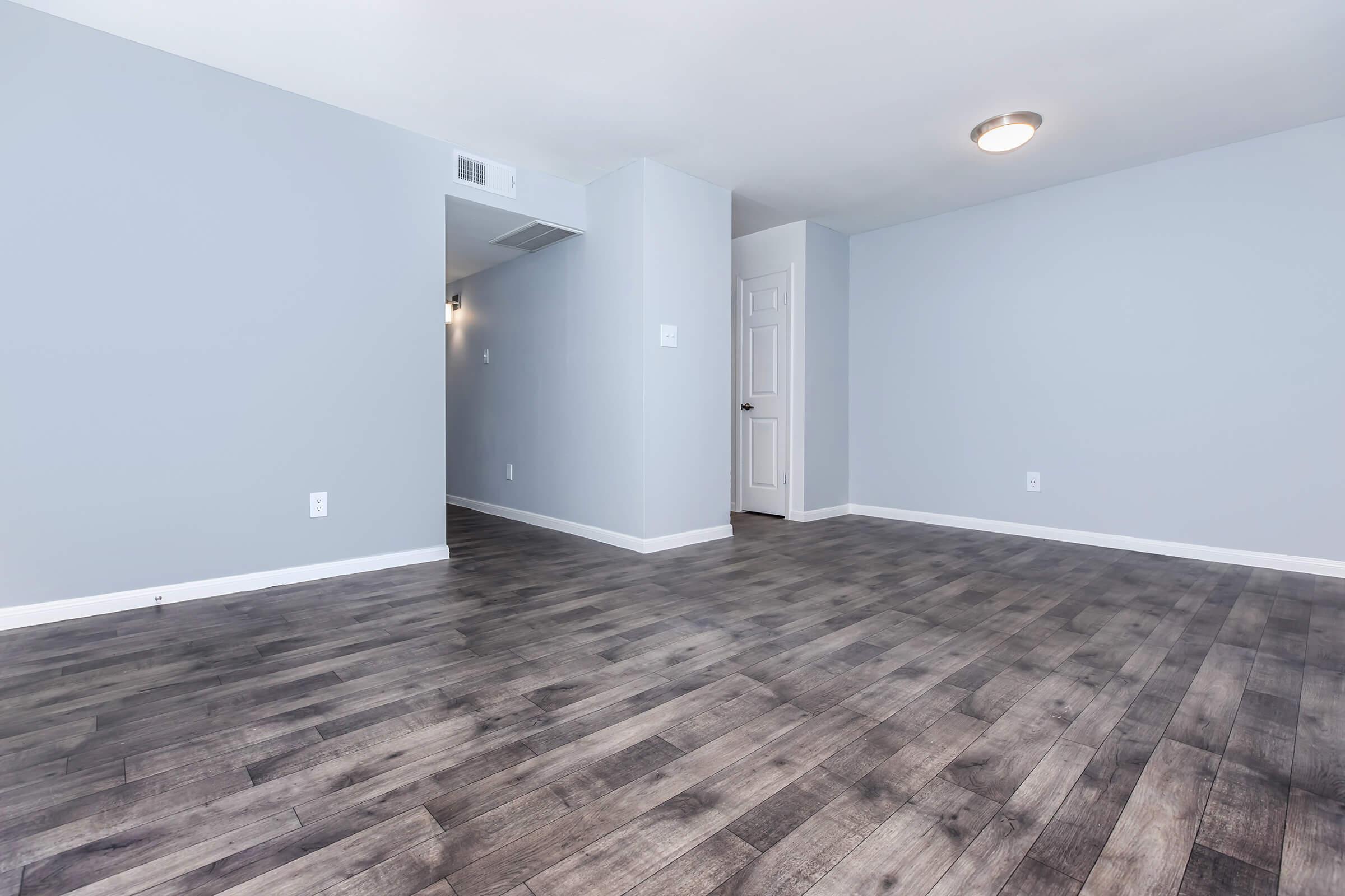 A bright, empty room with gray walls and dark wood-like laminate flooring. A single light fixture is mounted on the ceiling, and a closed door can be seen in the background. The space appears to be freshly renovated and is free of any furniture or decorations.