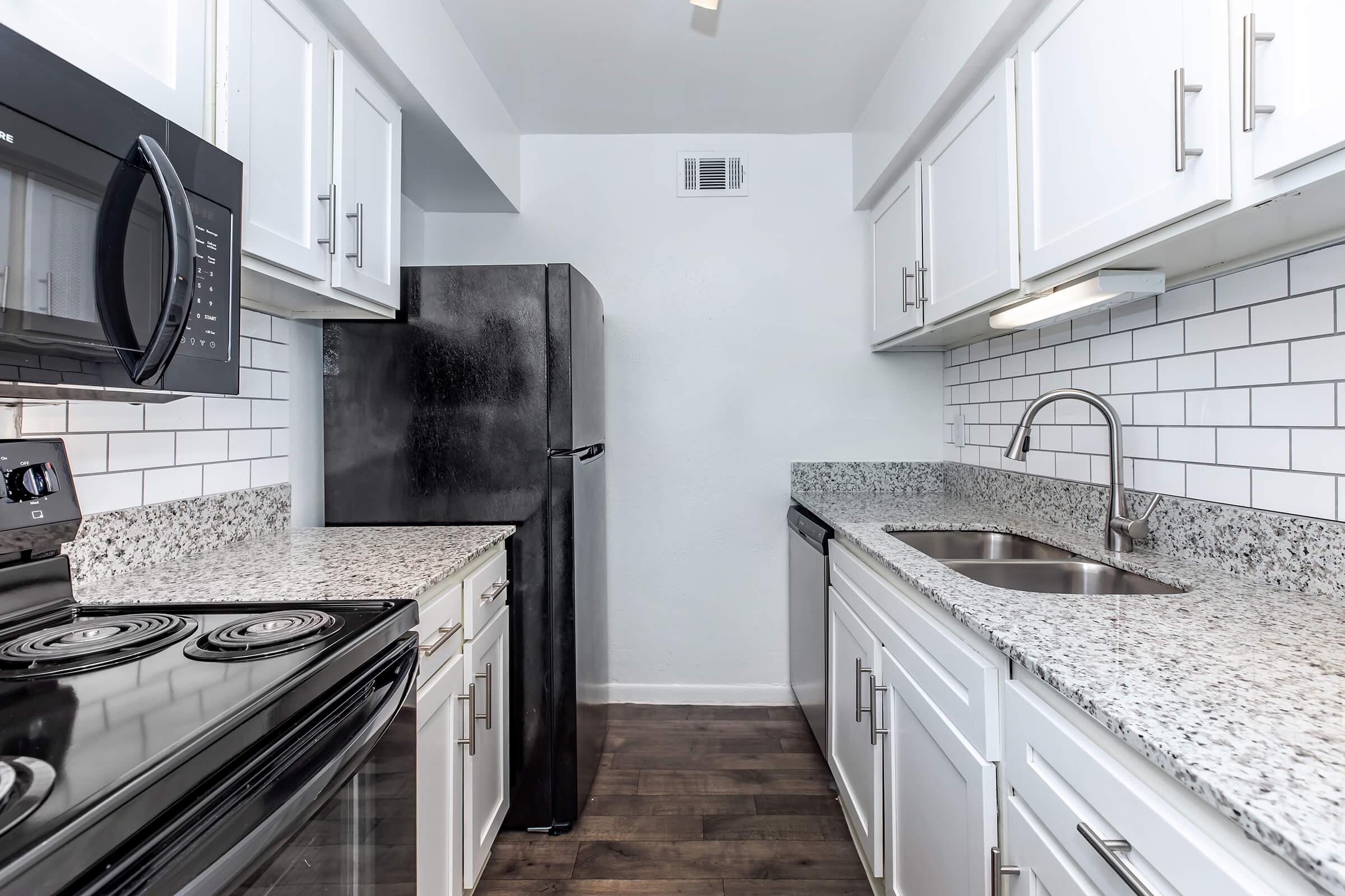 Modern kitchen featuring white cabinetry, granite countertops, and stainless steel appliances. The space includes a black refrigerator, a microwave above a stove, and a double sink with a sleek faucet. Subway tile backsplash adds a contemporary touch, and the flooring is dark wood for contrast.