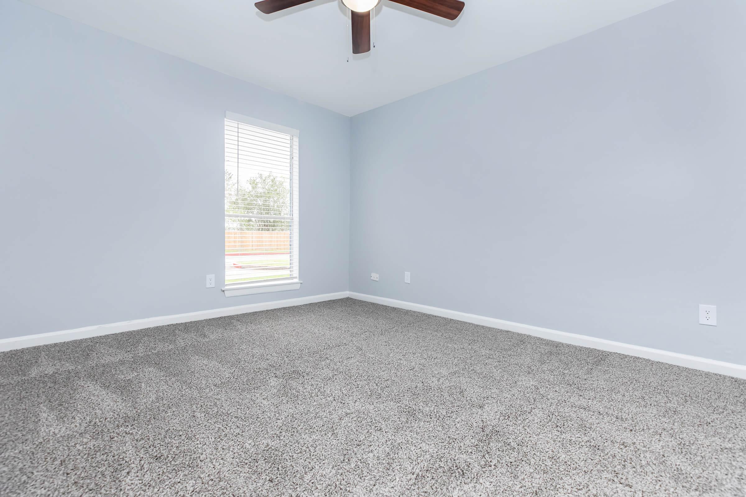 A lightly colored room with light gray walls and a ceiling fan. The floor is covered with plush, beige carpet. A window with blinds allows natural light to enter, illuminating the empty space. The room appears tidy and ready for furnishing, with no visible decorations or furniture.