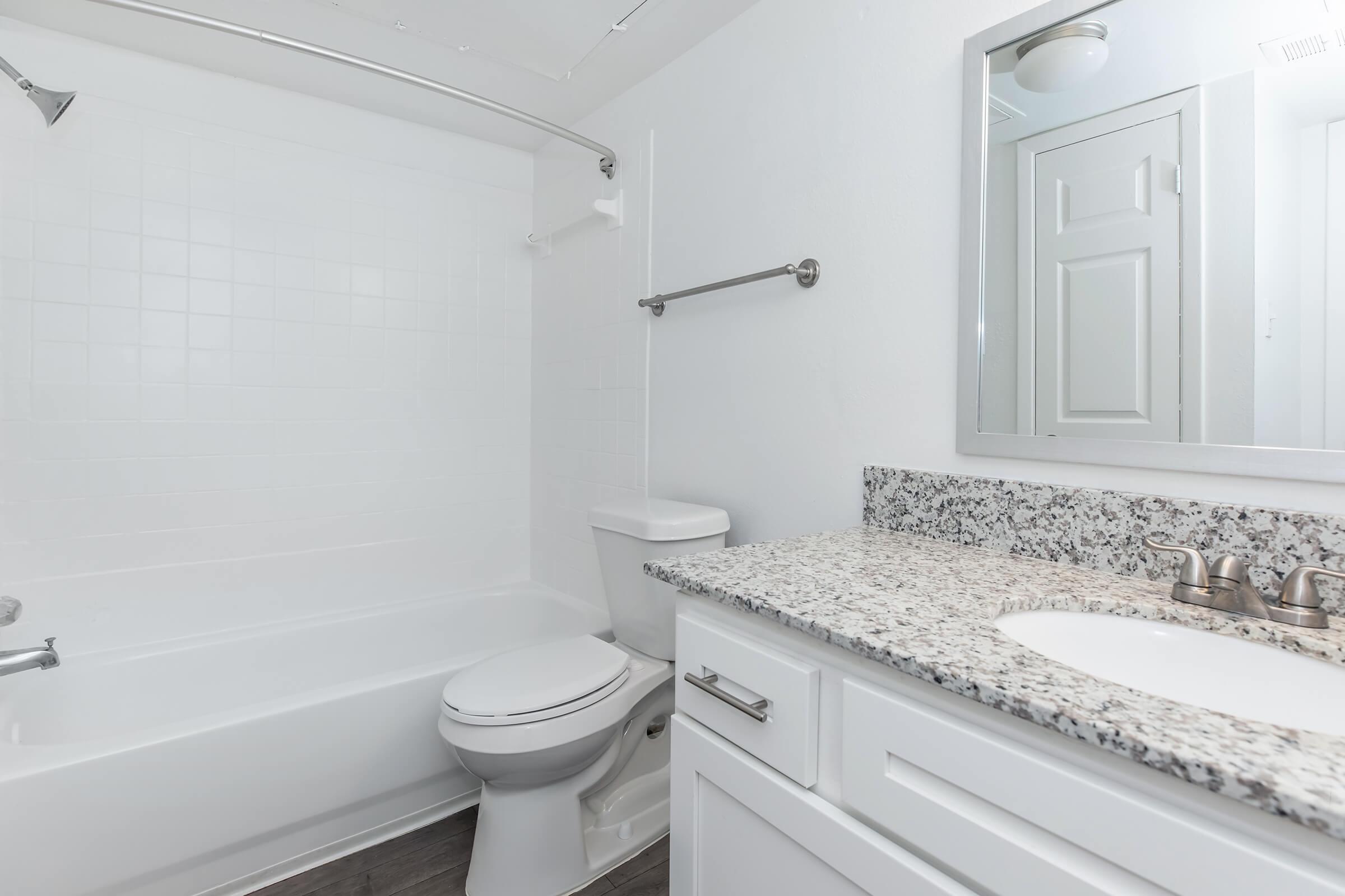 A clean, modern bathroom featuring a bathtub with a shower, a white toilet, and a vanity with a granite countertop. There is a round mirror above the sink, and the walls are painted white, creating a bright, airy atmosphere. A towel bar is mounted on the wall, and the floor is a dark wood finish.