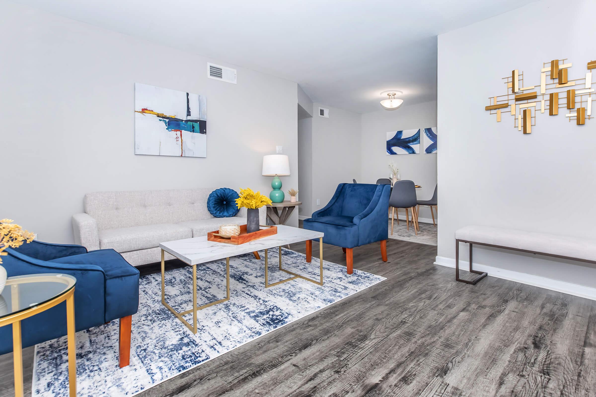 A modern living room featuring a light gray sofa and two blue armchairs, complemented by a decorative coffee table and a patterned rug. Wall art and a stylish lamp enhance the decor, while a dining area is visible in the background, showcasing a round table and chairs.