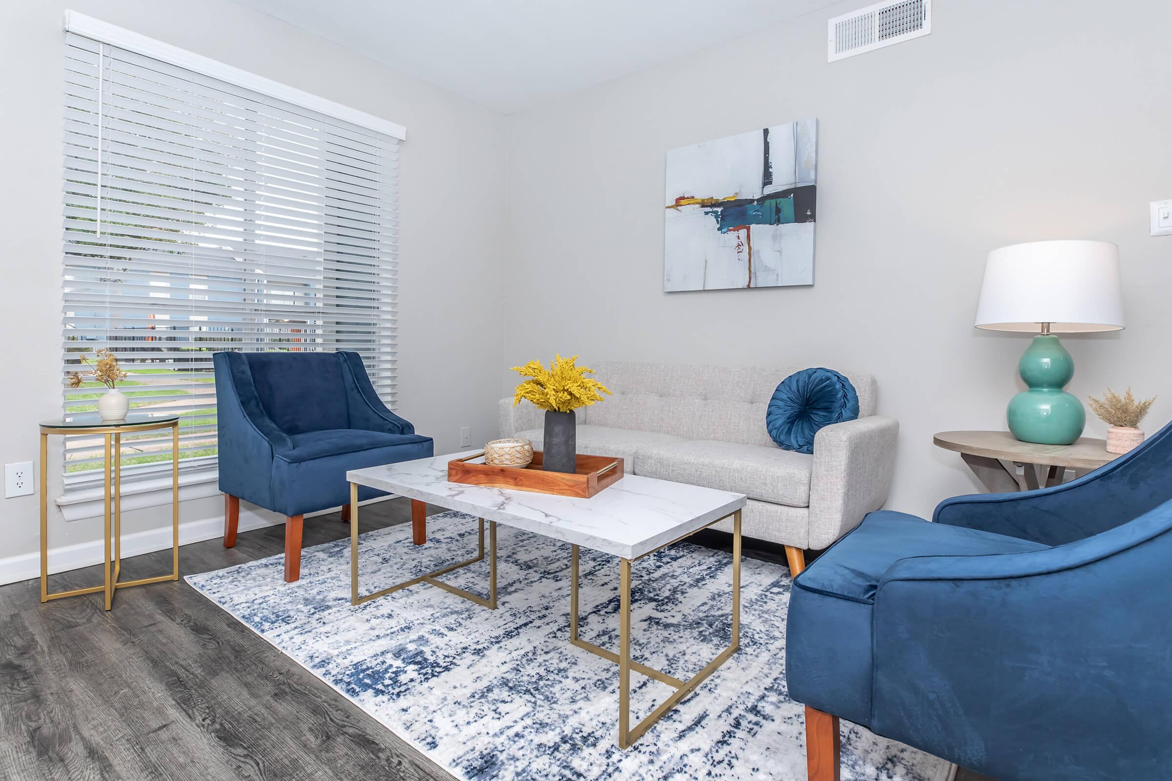 A modern living room featuring two blue armchairs and a light gray sofa. A marble-topped coffee table sits on a patterned blue and white rug. A decorative bowl and yellow flowers are on the table. Natural light streams in through blinds on the window, alongside a simple wall art piece.