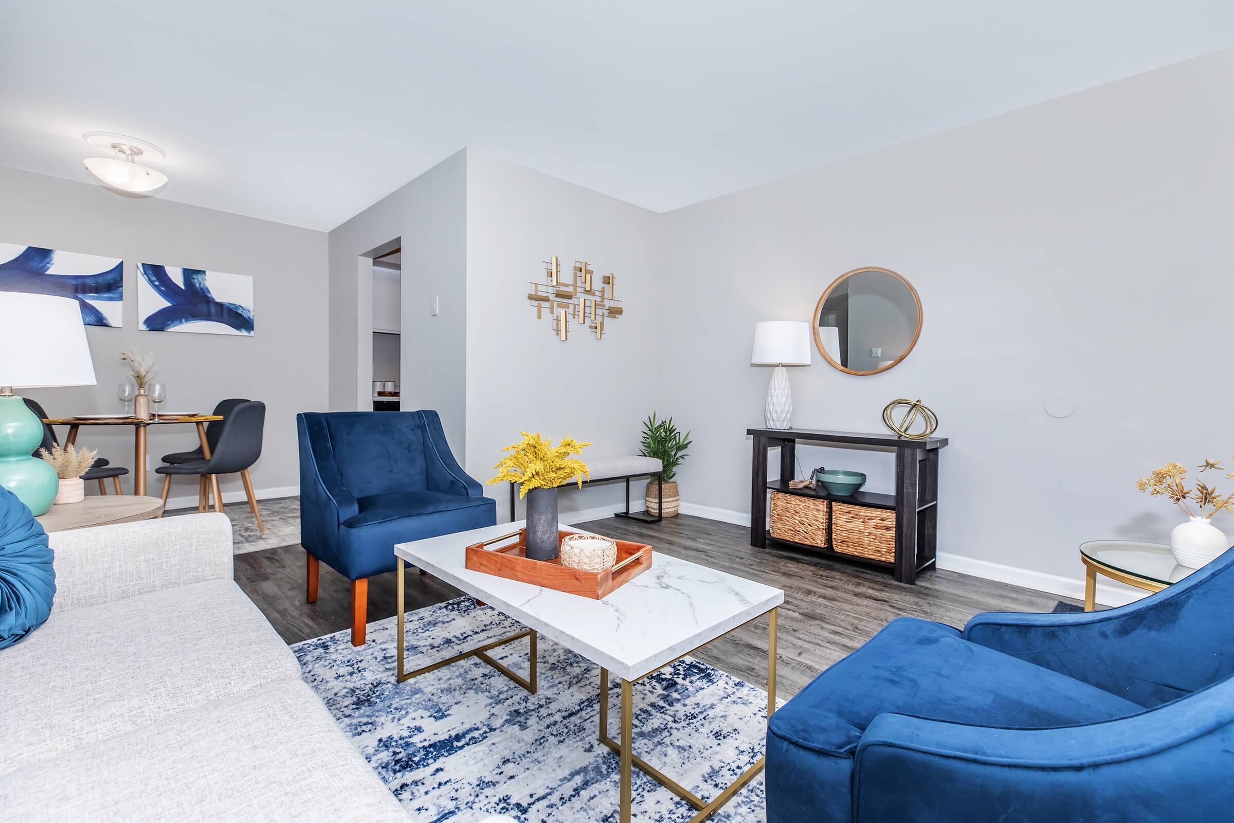 A modern living room featuring a light-colored sofa, two blue accent chairs, a marble coffee table, and decorative elements like a round mirror, a lamp, and a plant. The space is styled with artwork on the wall and a dining area visible in the background.
