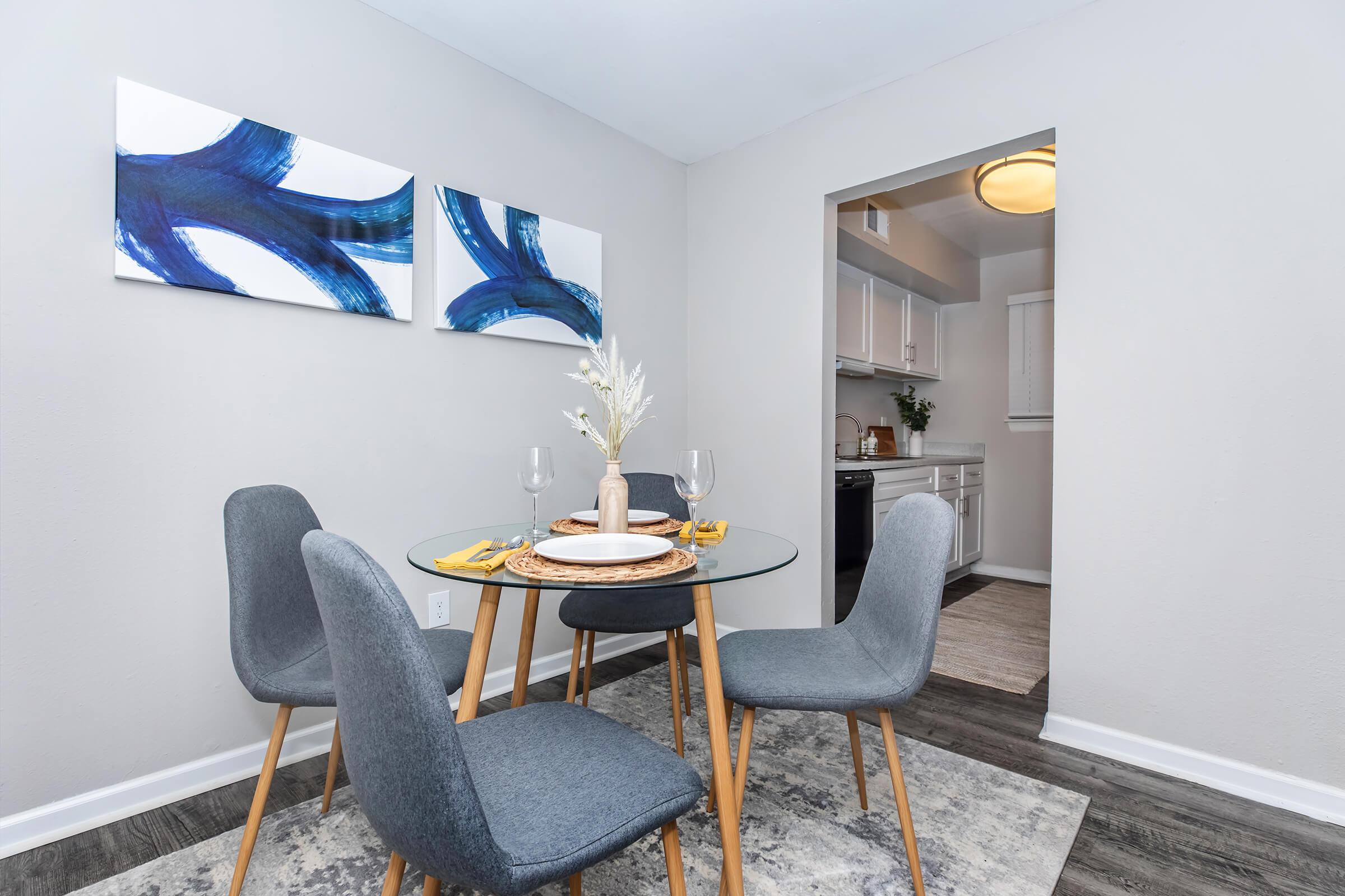 A modern dining area featuring a round glass table set with plates and glasses, surrounded by four gray chairs. Two abstract blue paintings adorn the wall, while a doorway leads to a kitchen area in the background. A light rug enhances the space, adding a cozy feel.