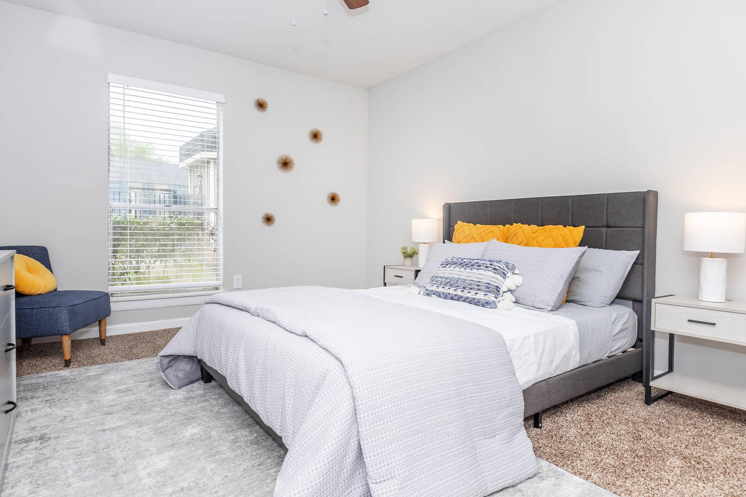 A cozy, modern bedroom featuring a grey upholstered bed with white and yellow bedding, a small blue armchair, two bedside tables with lamps, and a window with white blinds offering natural light. Decorative wall accents are visible, and the room has soft, neutral tones with a plush, light-colored carpet.