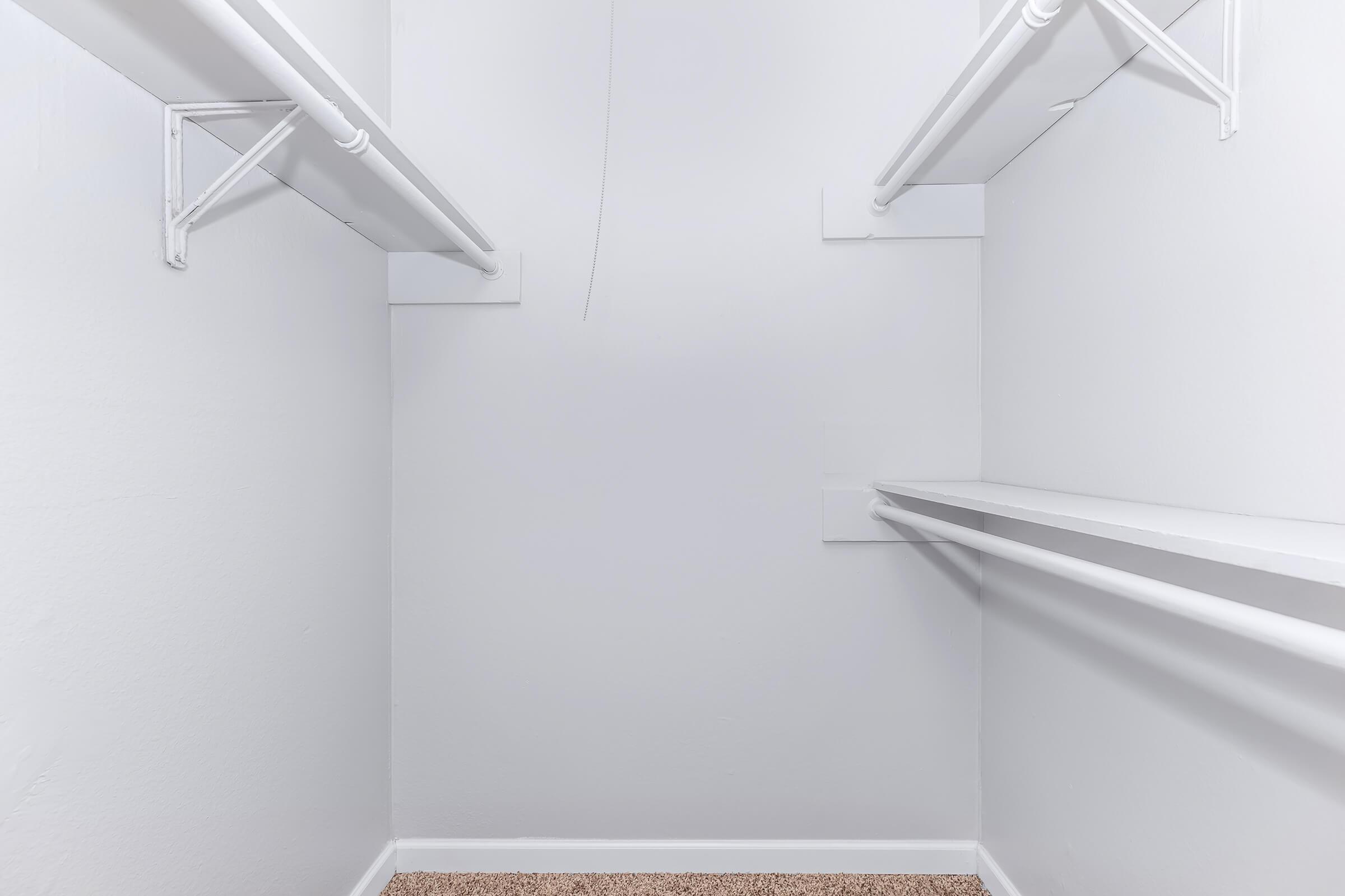 Empty closet interior with white walls, featuring two horizontal shelves and two hanging rods installed on opposite sides. The floor has a light carpet, and there is a minimalistic, uncluttered appearance.