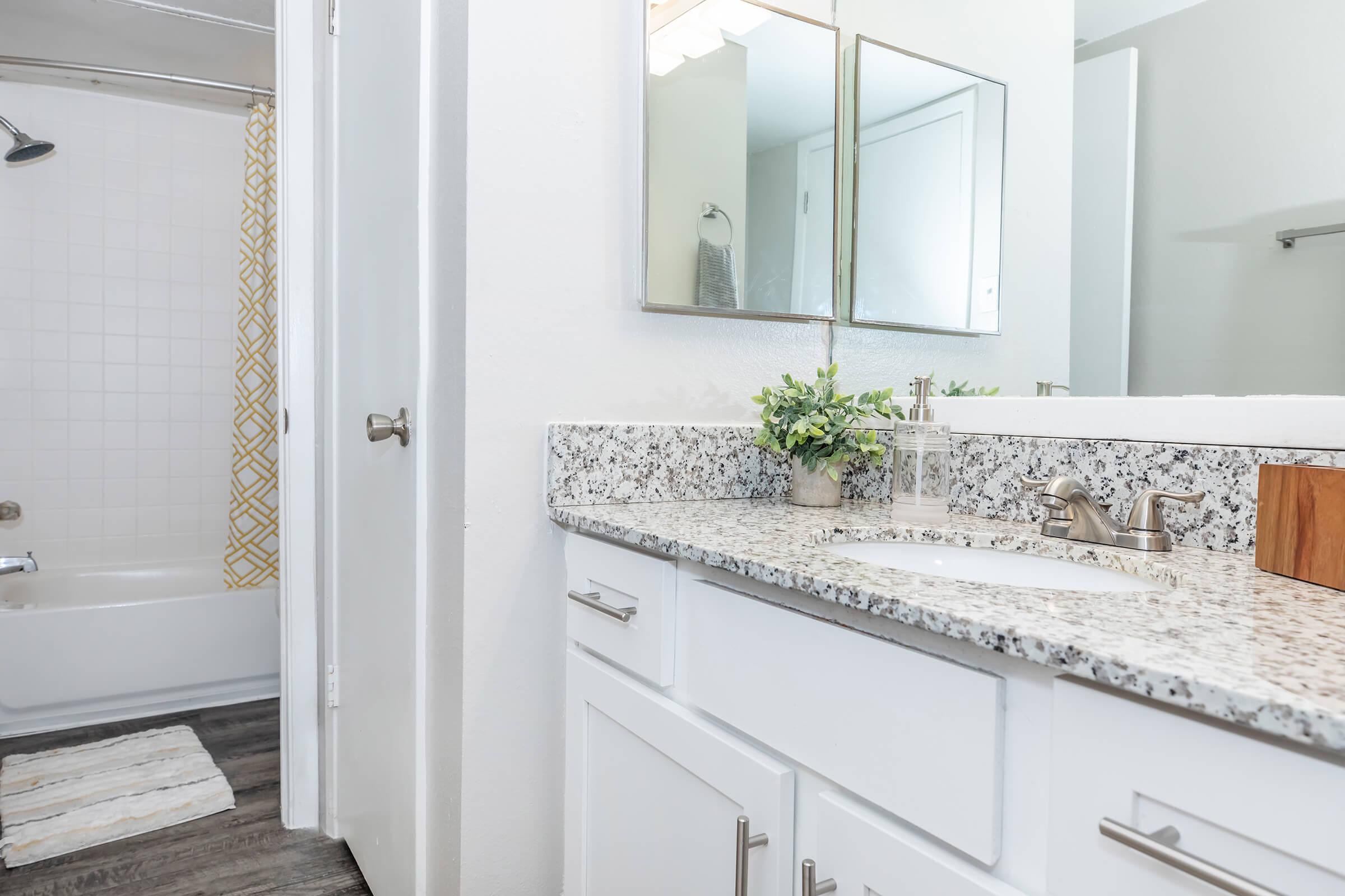 A modern bathroom featuring a white countertop with a sink, stylish faucets, and decorative plants. There is a shower and tub with a beige and yellow patterned shower curtain visible in the background. The space has a clean, minimalist design with neutral colors and wood-like flooring.