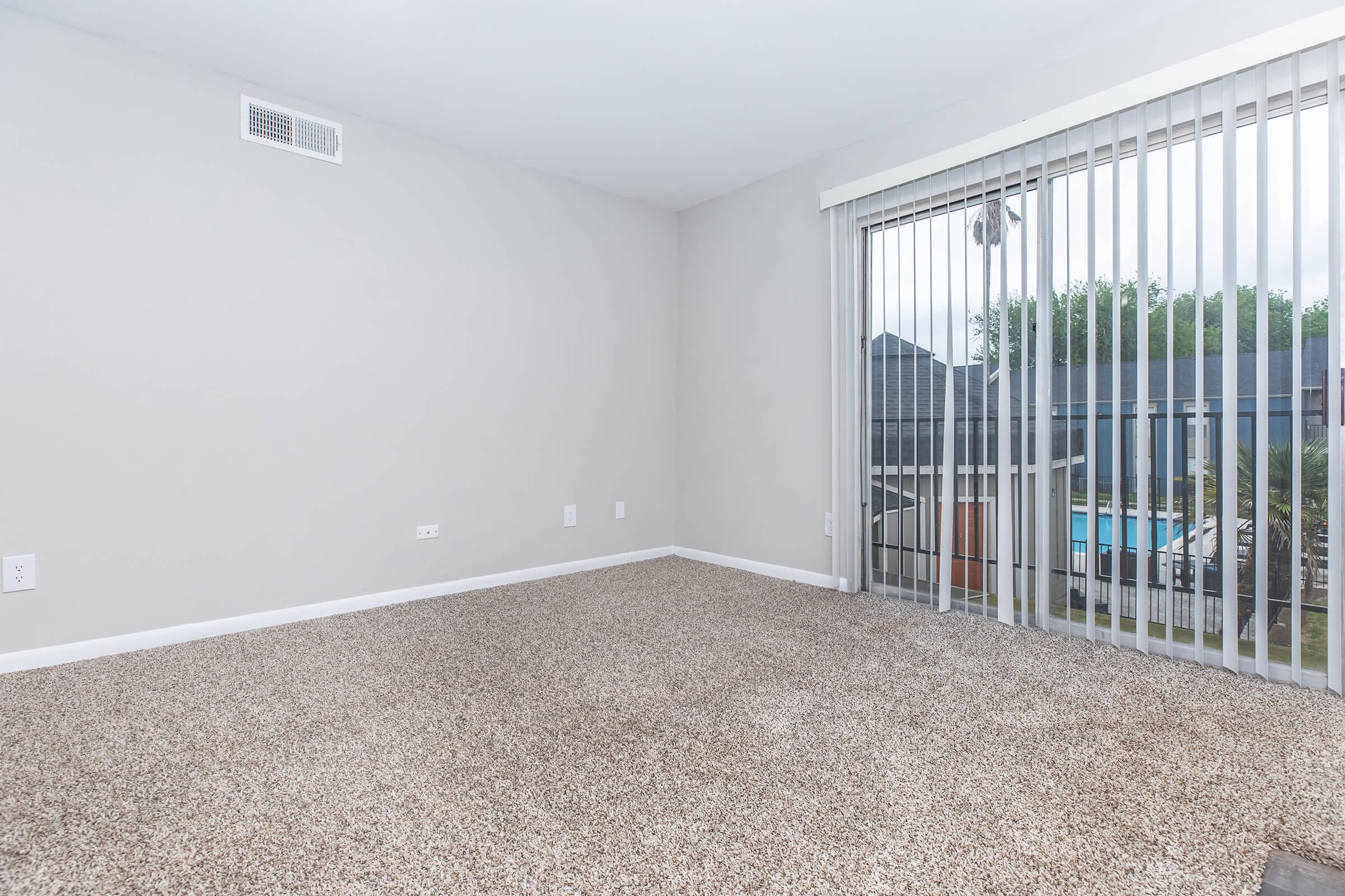 An empty room with light gray walls and beige carpeting. There is a large sliding glass door with vertical blinds that leads to a patio area. The view outside shows trees and a glimpse of a swimming pool.