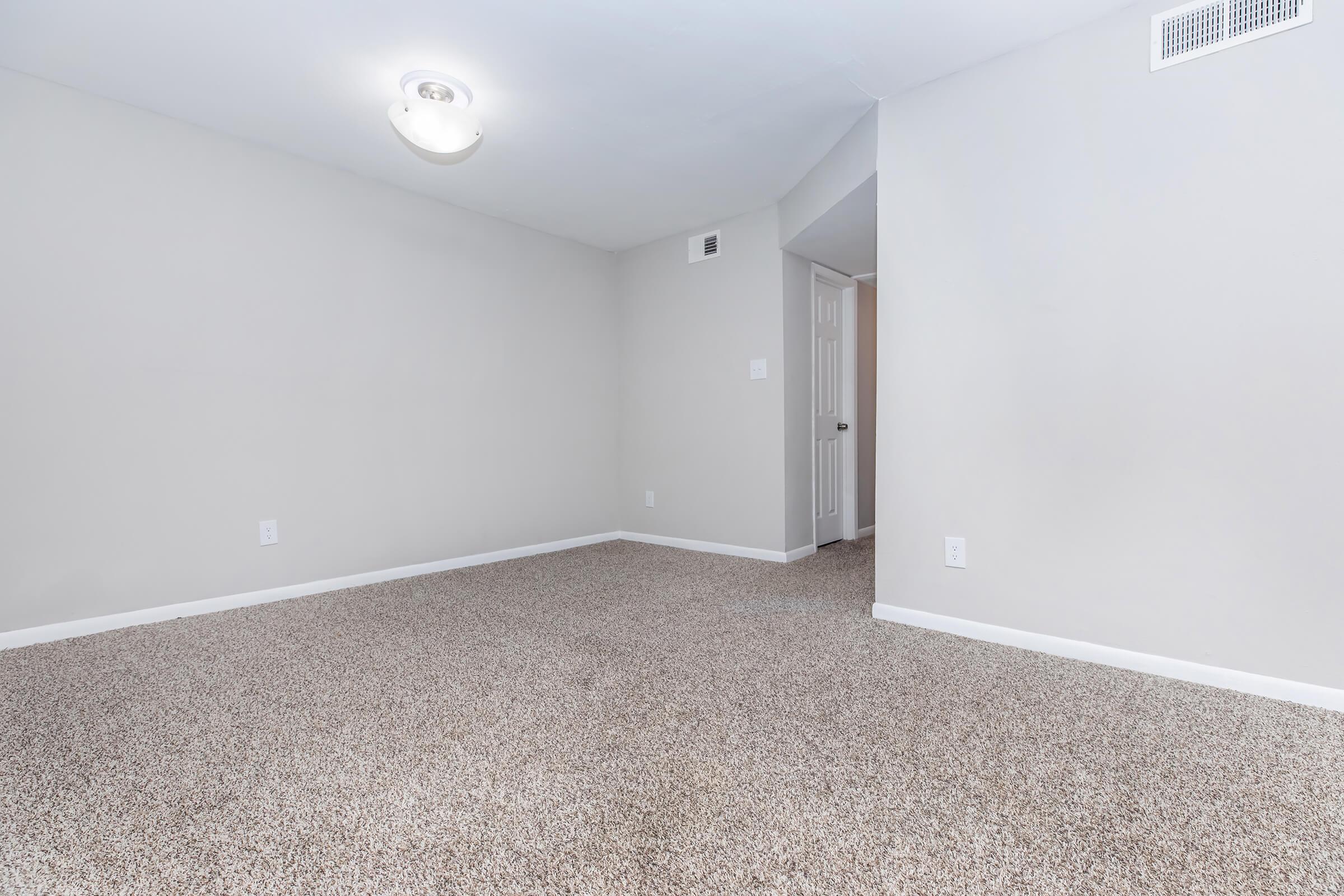 Empty room with light gray walls and carpeted floor. A ceiling light fixture is visible, and there is a doorway leading to another room on the right. The space is clean and uncluttered, with no furniture present.